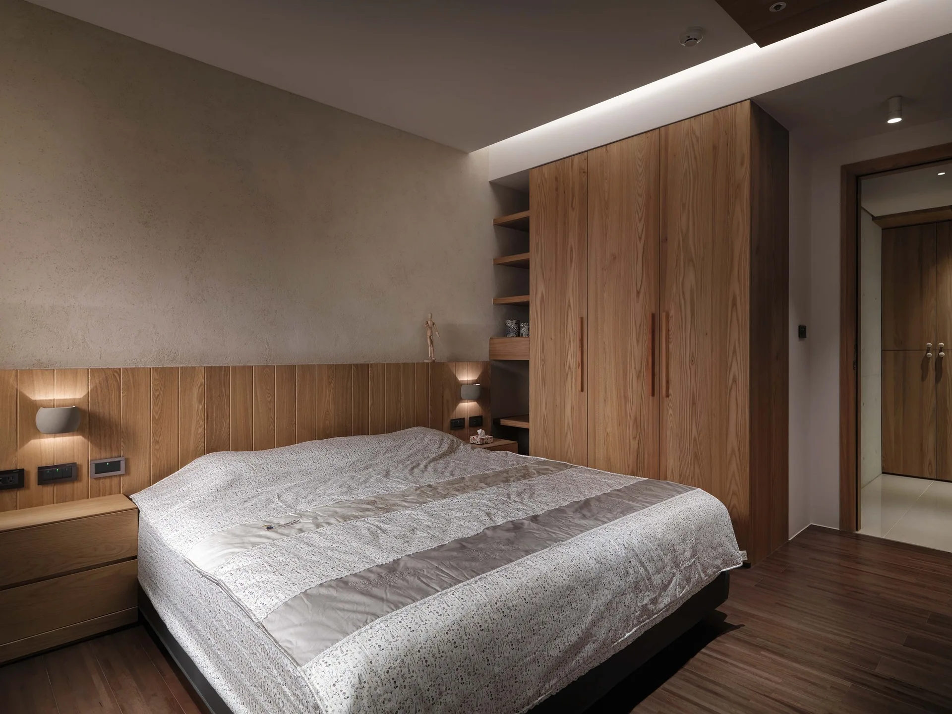 A minimalist bedroom featuring a bed with patterned sheets, a built-in wooden wardrobe, lower wall wood paneling, and warm cove lighting.