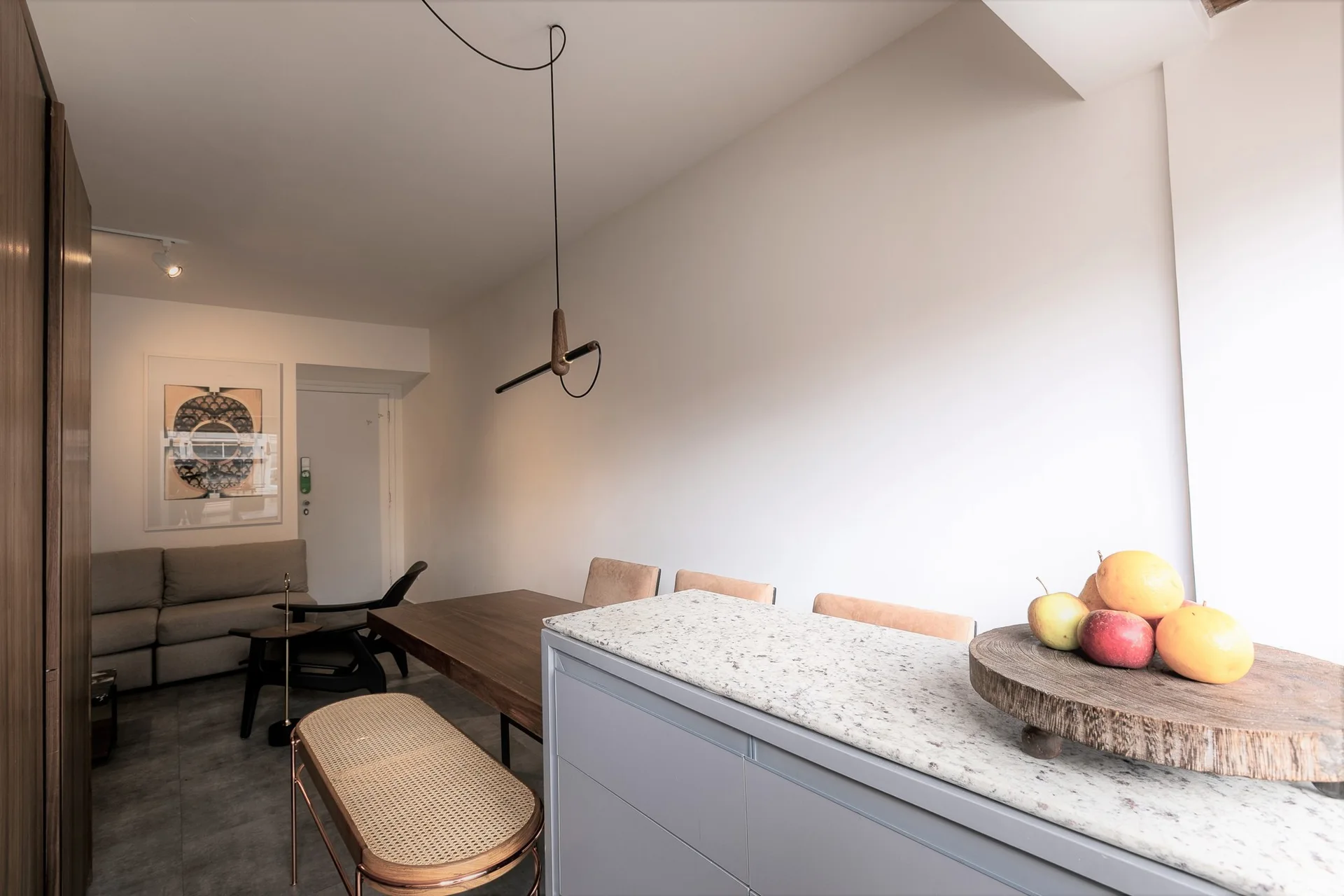 A minimalist dining area featuring a wooden table, a rattan-topped bench, and a grey kitchen counter with a speckled stone surface.