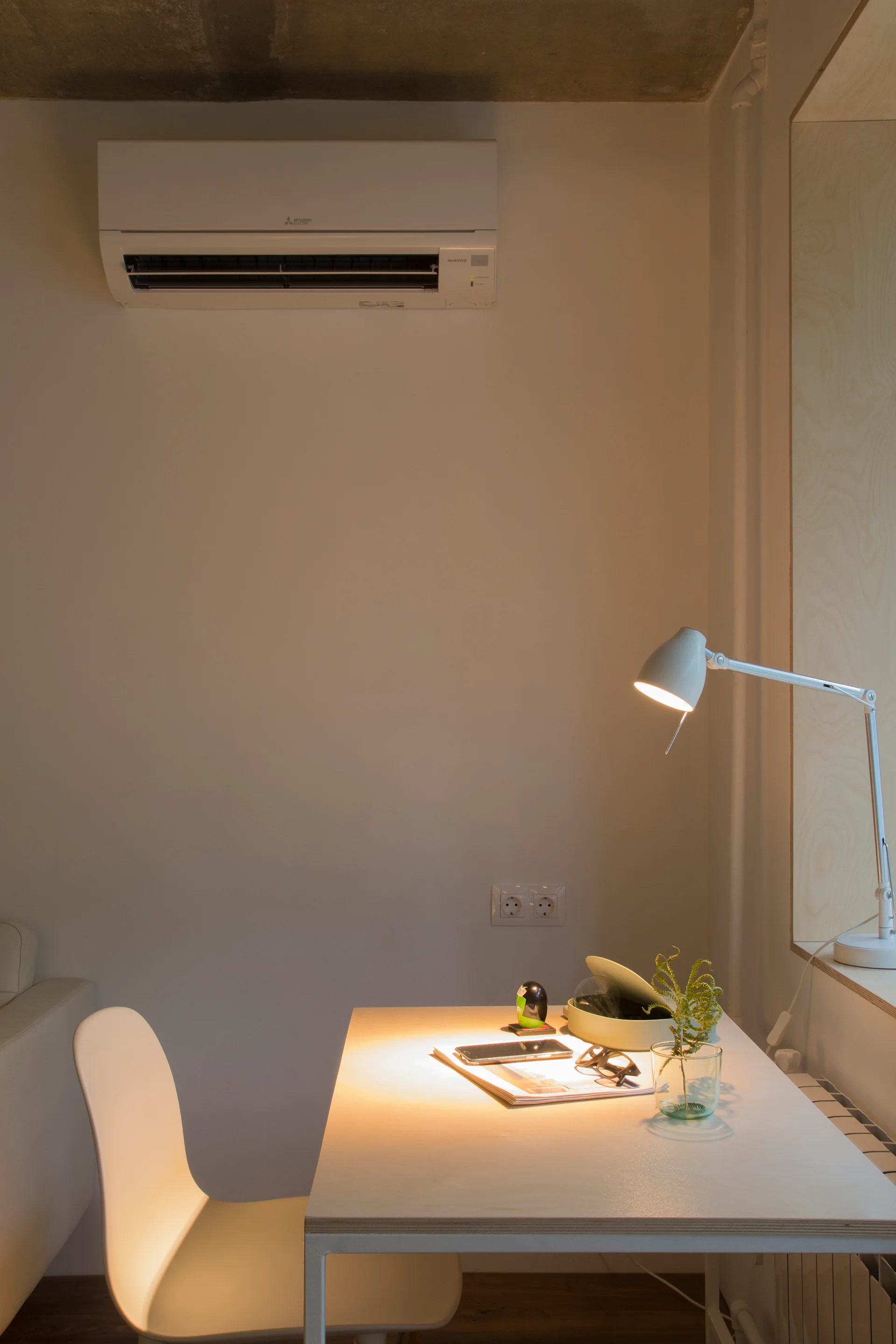A modern white desk and chair sit beneath a white wall-mounted air conditioner, lit by a warm task lamp near a light wood window frame.