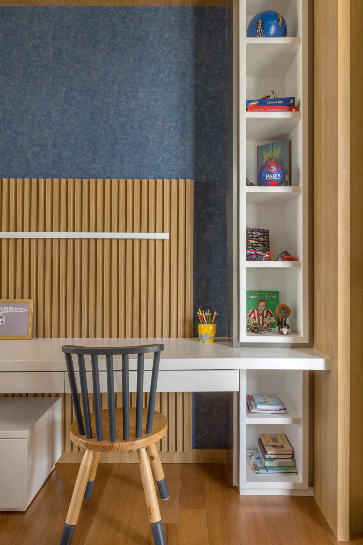A kids study desk with a spindle-back wooden chair, set against a vertical wood slat wall accent and built-in white shelving displaying toys.