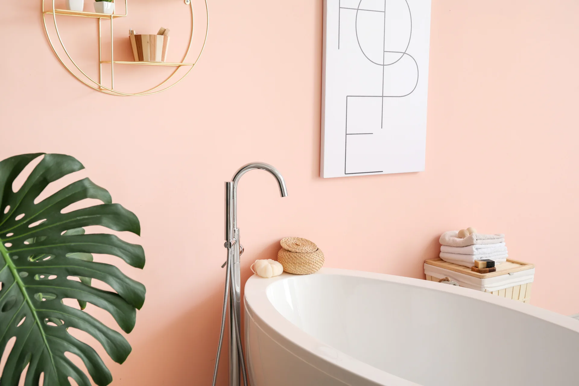 A white freestanding bathtub stands against a soft pink wall with a chrome floor faucet, a round gold shelf, and a green monstera leaf.