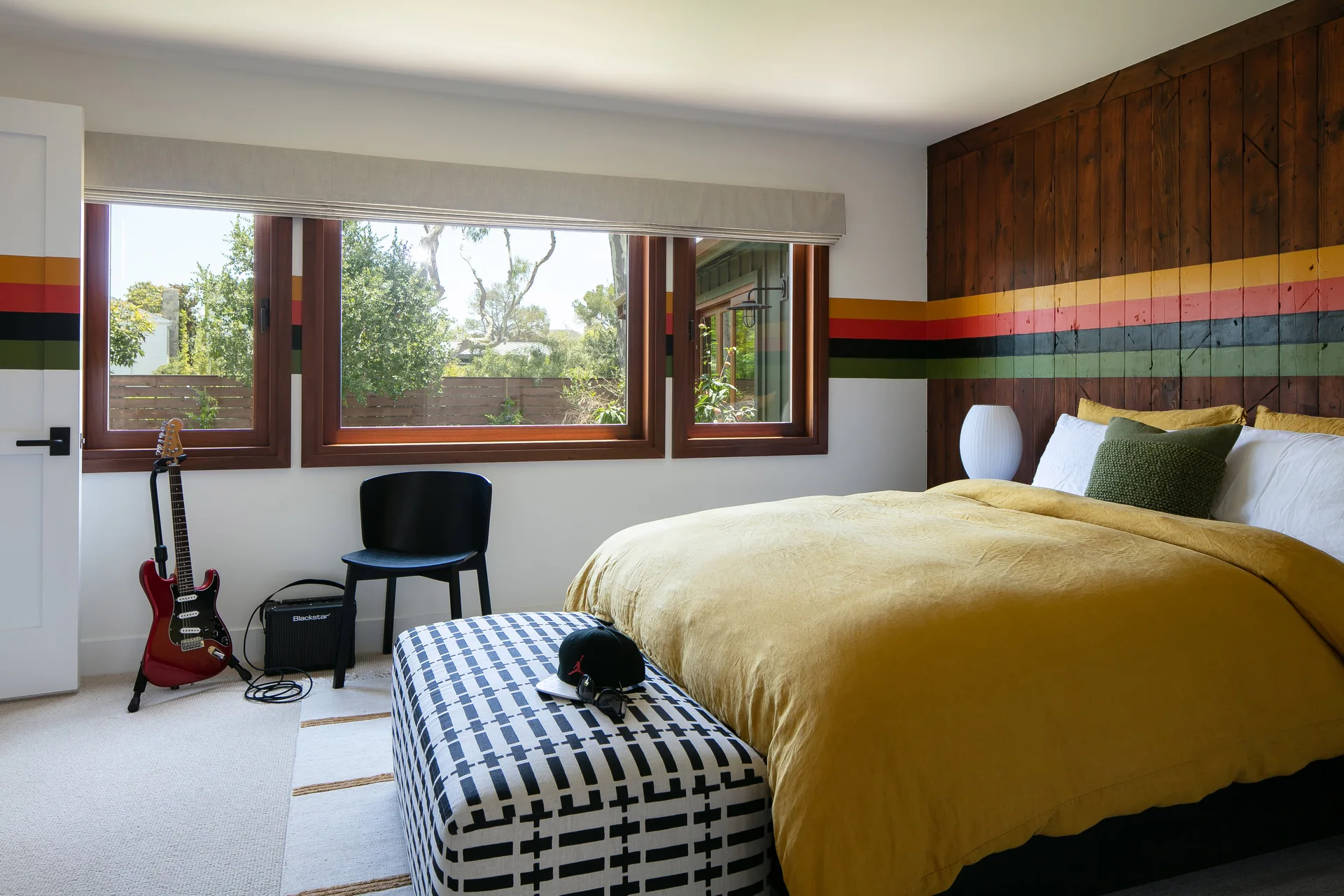 A bedroom features a bed with a yellow duvet, a black-and-white grid pattern bench, and a wood-paneled accent wall with horizontal stripes.