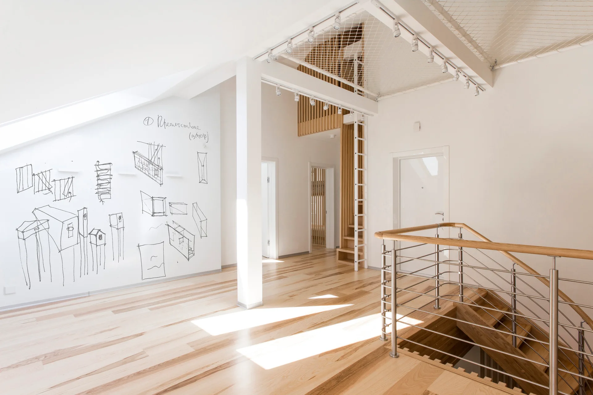 A bright loft hallway features light wood floors, a metal stair railing, interior doors, and hand-drawn black sketches on a slanted white wall.