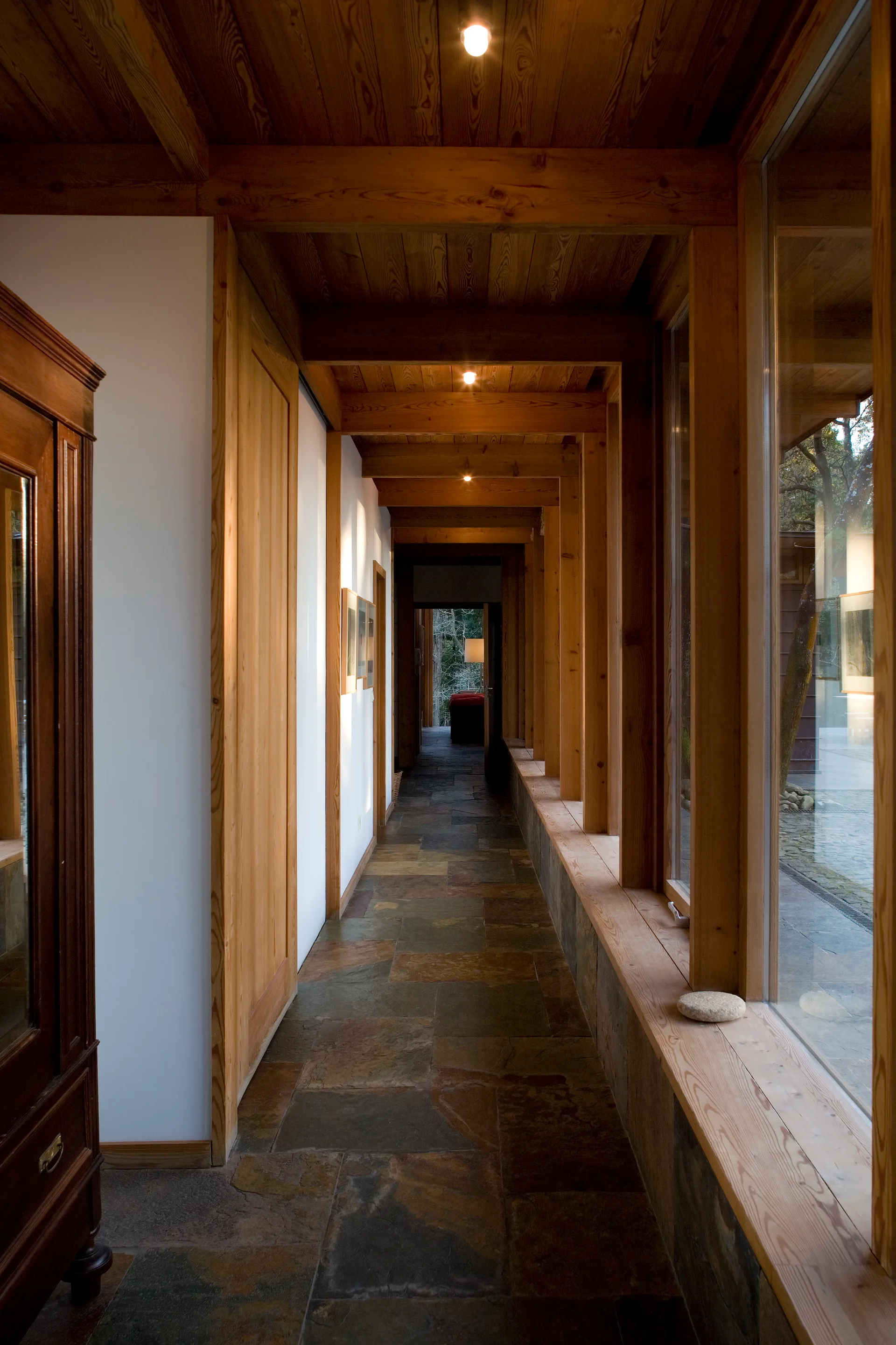 A long hallway features a slate stone tile floor, exposed wood ceiling beams, white walls, and floor-to-ceiling windows with wood frames.