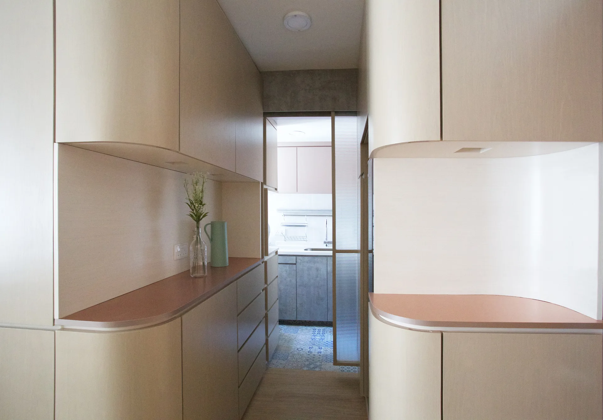 A narrow hallway with custom light wood cabinetry with rounded edges leading to a kitchen behind a ribbed glass sliding door.