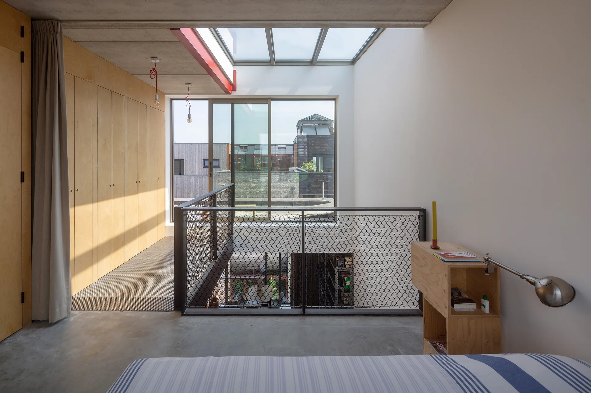 A modern mezzanine landing features a plywood wardrobe, polished concrete floor, and black metal mesh railing under a large bright skylight.