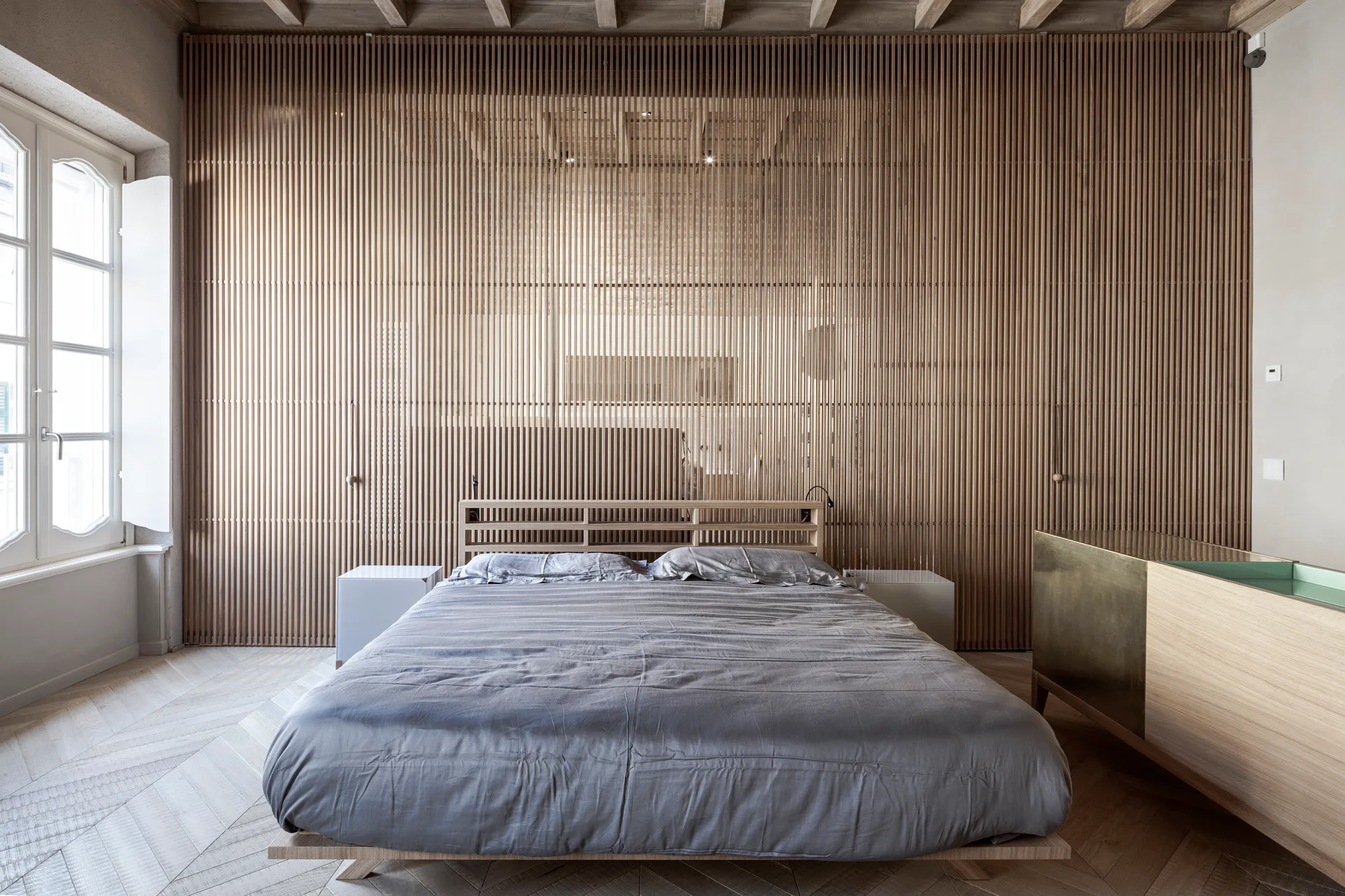 A minimalist bedroom features a large vertical wood slat wall behind a low bed with grey linens and light wood chevron flooring.