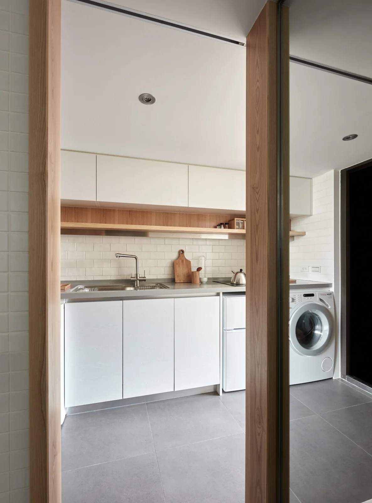 A minimalist white kitchen featuring a subway tile backsplash, an integrated washing machine, and grey tile floors seen through a wood-trimmed doorway.