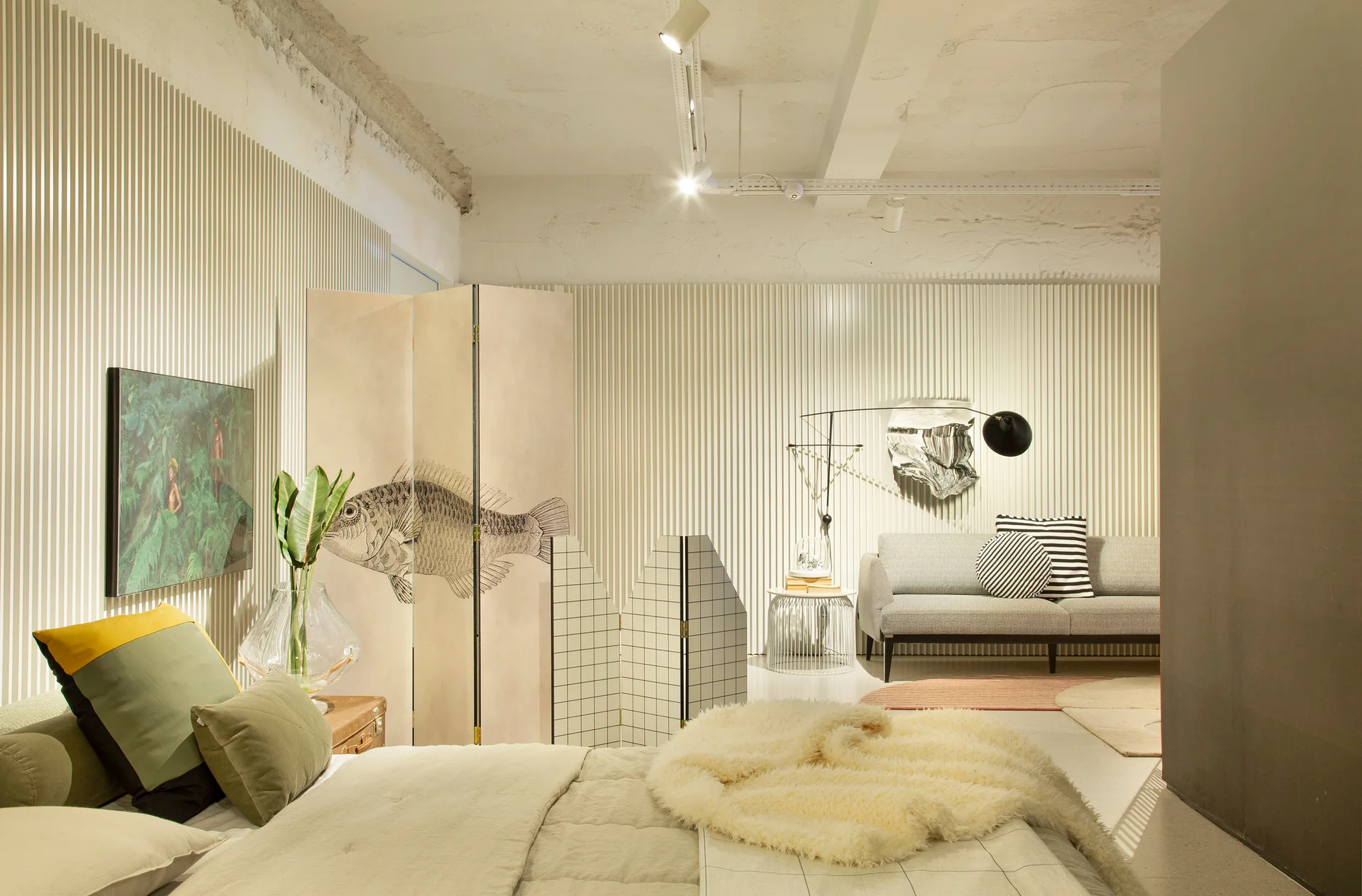 A contemporary bedroom features textured cream slatted walls, a bed with neutral linens, a decorative folding screen, and a grey sofa in the background.