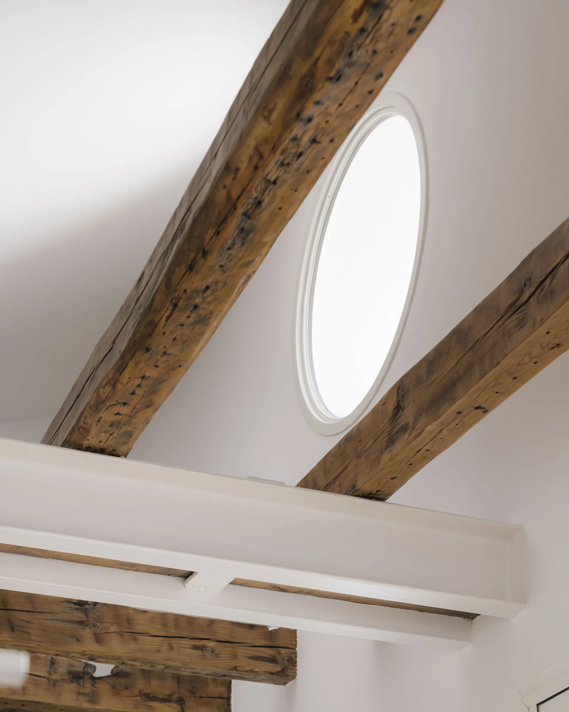 Exposed rustic wooden ceiling beams lie above a painted white steel I-beam and a round window set in a white wall.