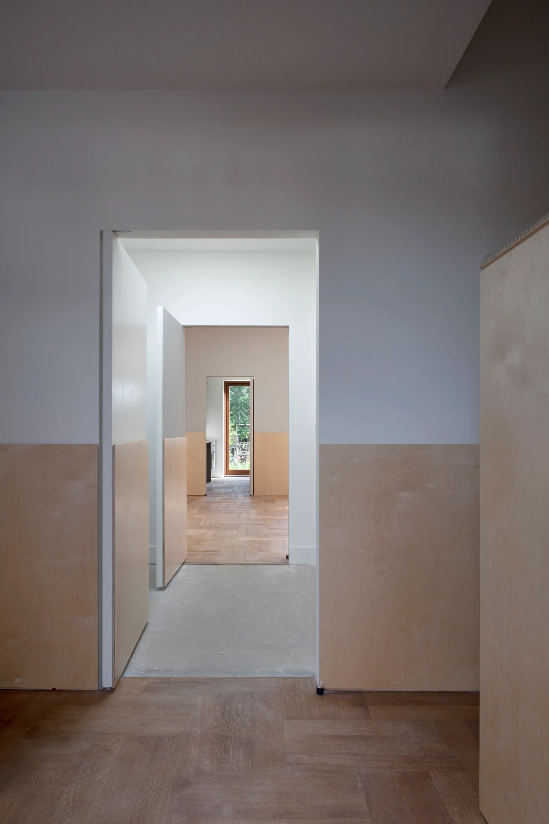 A perspective view down a minimalist hallway showing multiple open doorways, light wood wainscoting on white walls, and parquet flooring.
