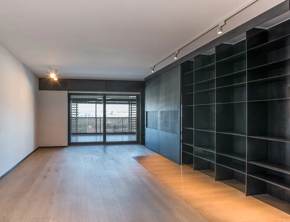 A minimalist room features light wood plank flooring, a large black metal grid shelving unit on one wall, and sliding glass balcony doors.