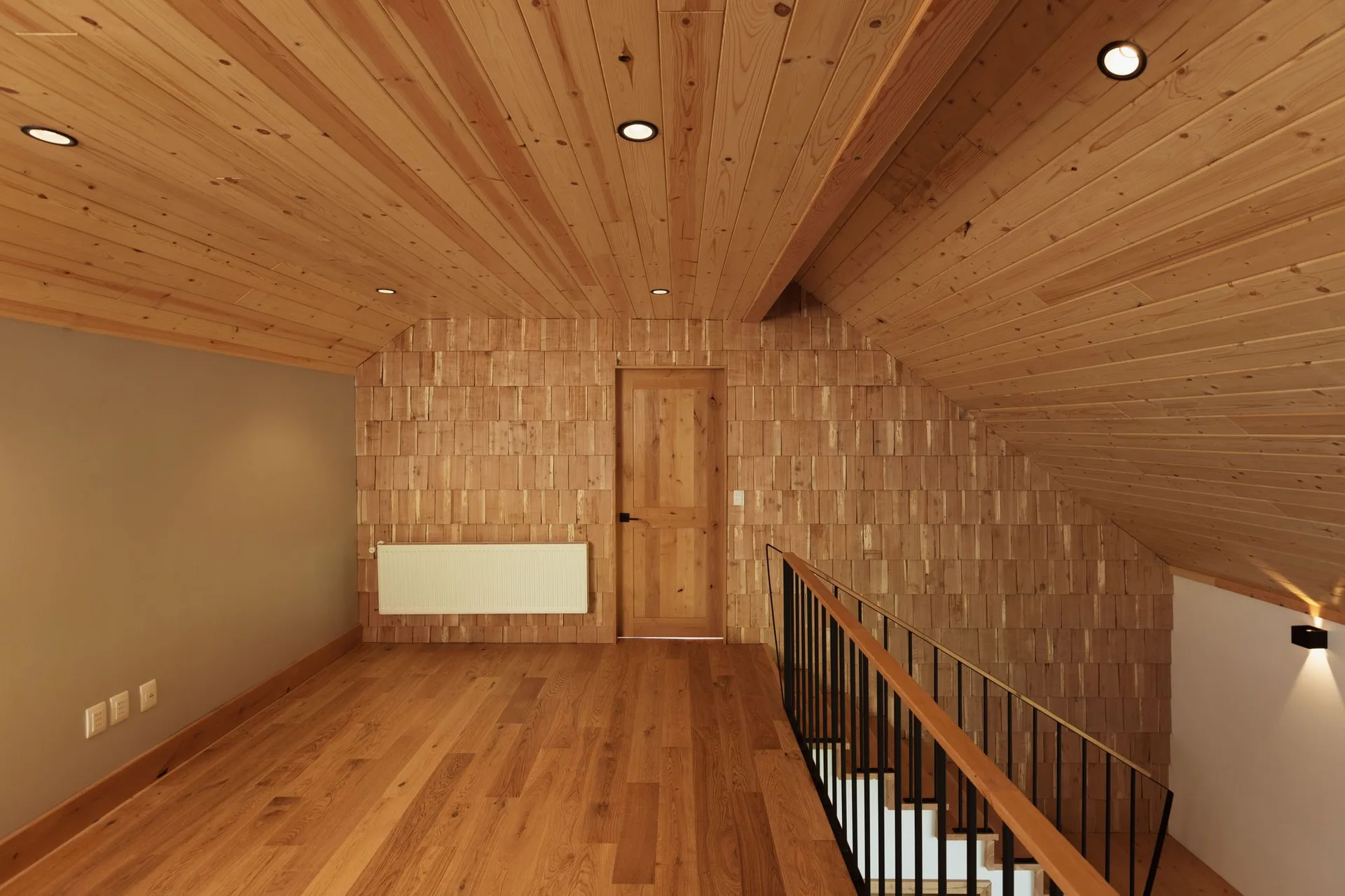 A residential attic landing featuring a wood-planked sloped ceiling, a shingled accent wall with a wooden door, and a black staircase railing.