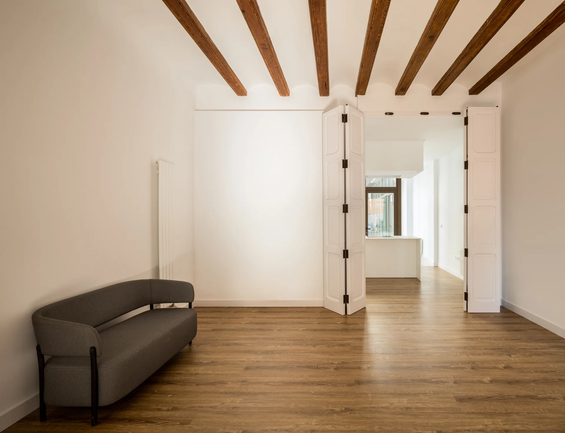 A minimalist room with a grey curved sofa, exposed wooden ceiling beams, medium wood flooring, and tall white folding doors leading to a kitchen.