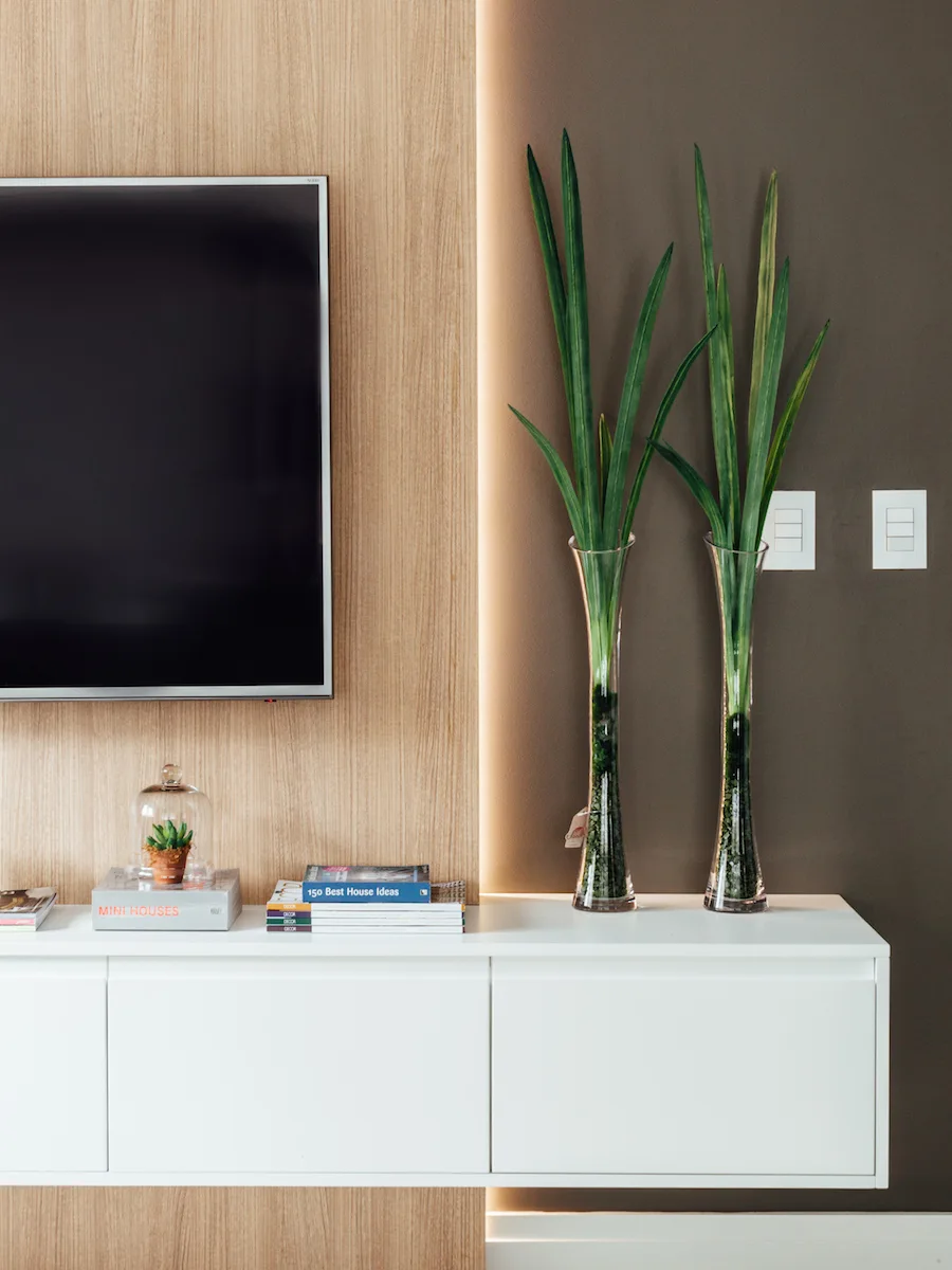A wall-mounted white media console features books and two tall glass vases under a flat-screen television mounted on a backlit wood panel.