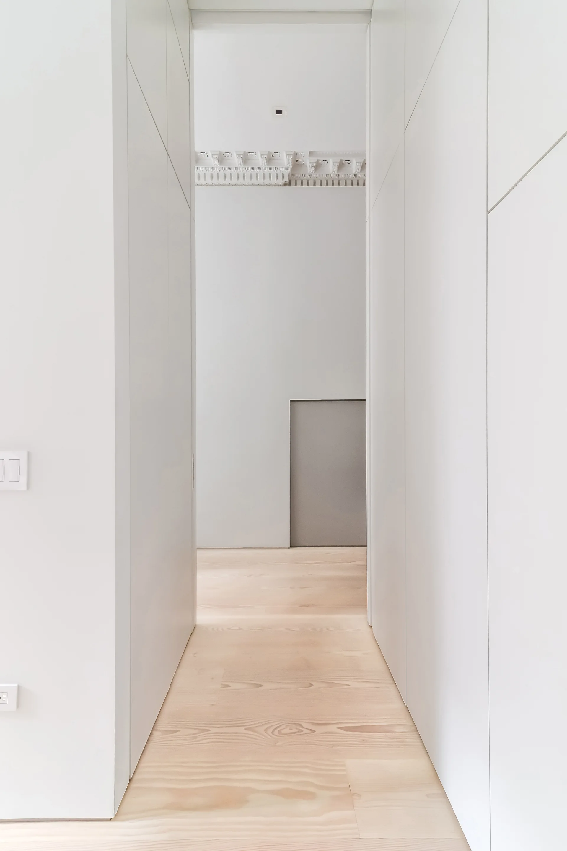 A minimalist hallway features white floor-to-ceiling cabinetry, light wood floorboards, and a view of an ornate plaster ceiling molding in the background.
