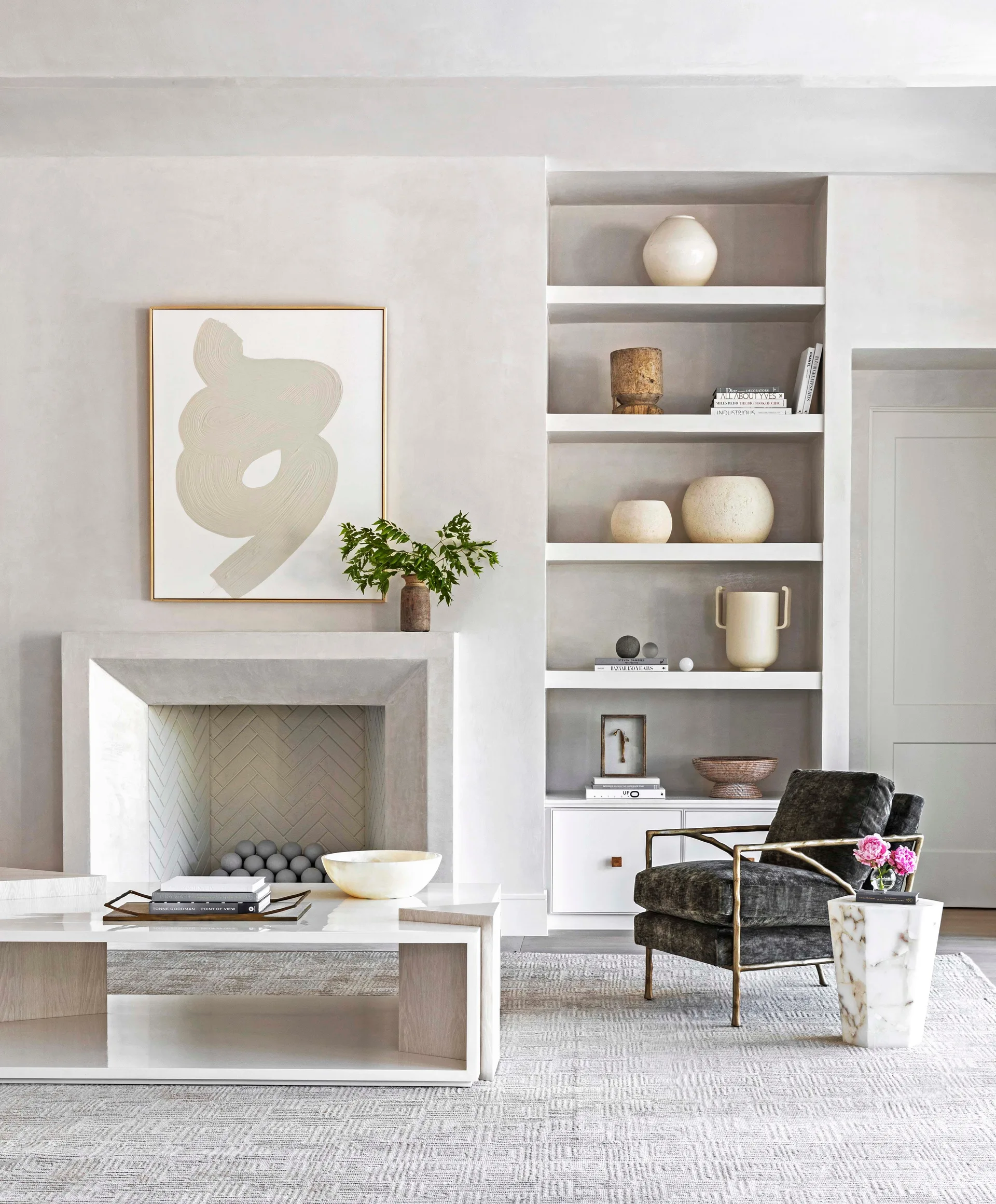 A neutral living room features a plaster fireplace with abstract art, built-in shelving with vases, a dark armchair, and a modern white coffee table.