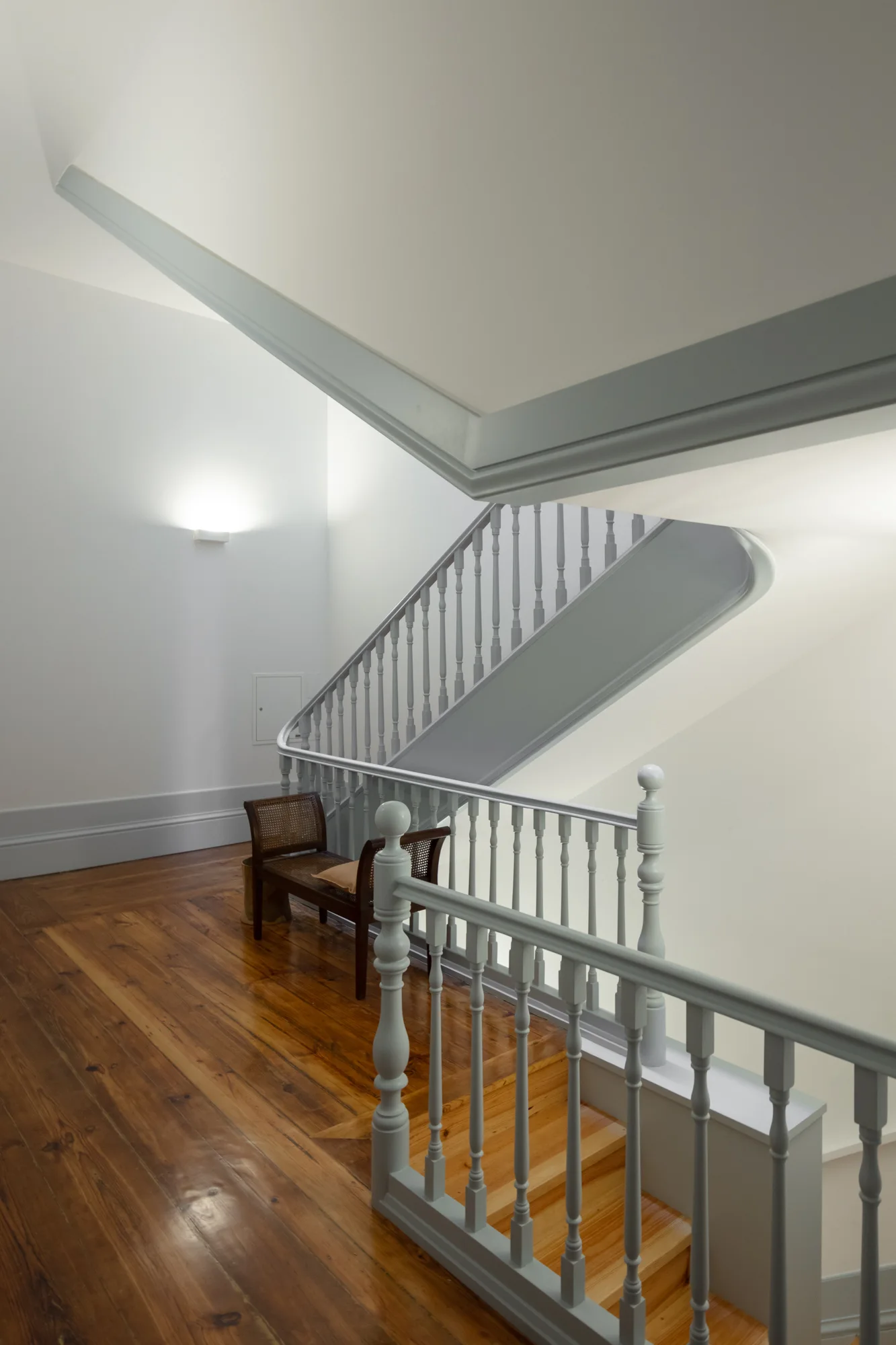 A staircase landing features a curved grey wooden railing, white walls, a varnished hardwood floor, and a cane bench.