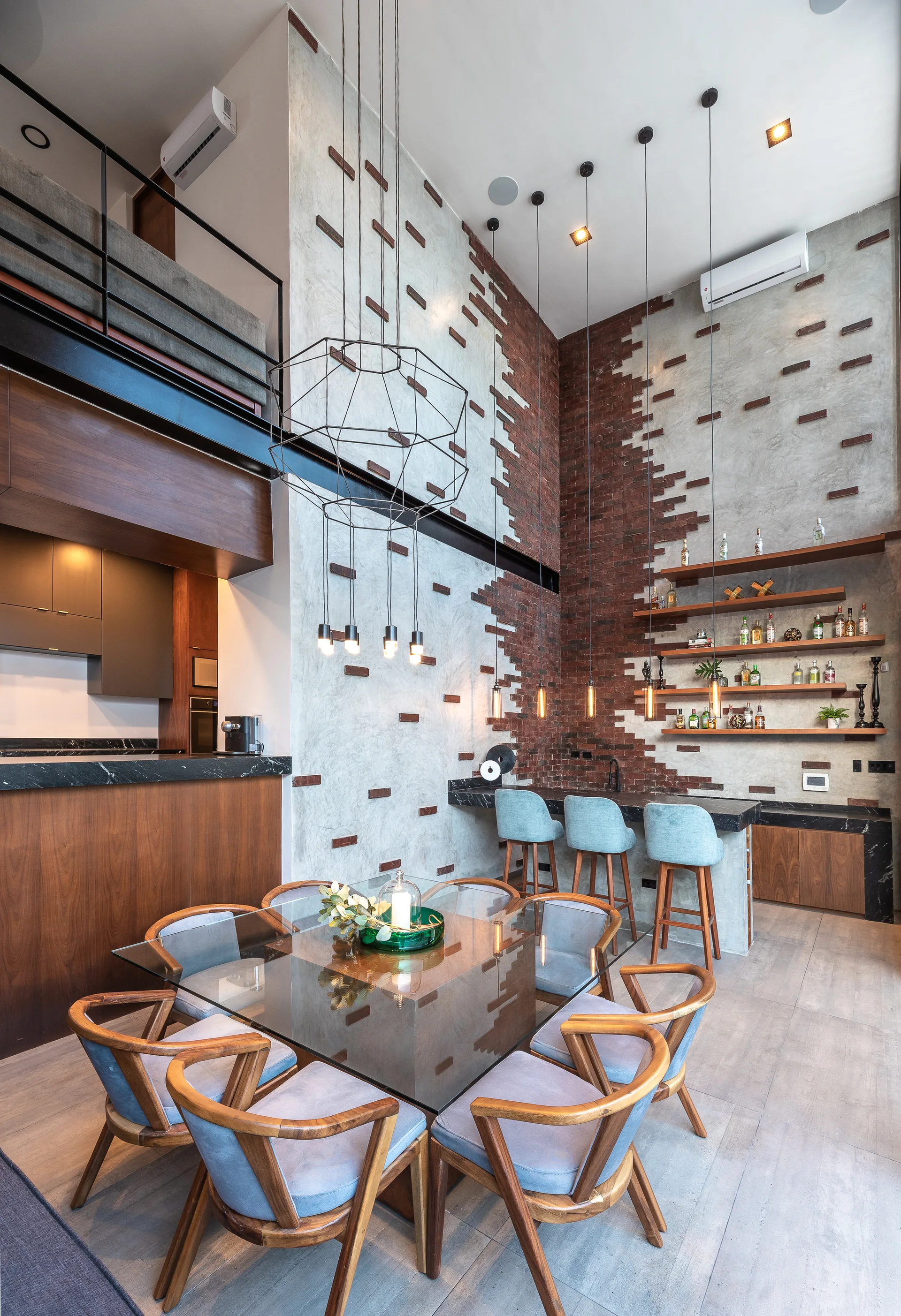 A double-height loft dining room features a glass table with blue chairs next to a home bar with an exposed brick and concrete accent wall.