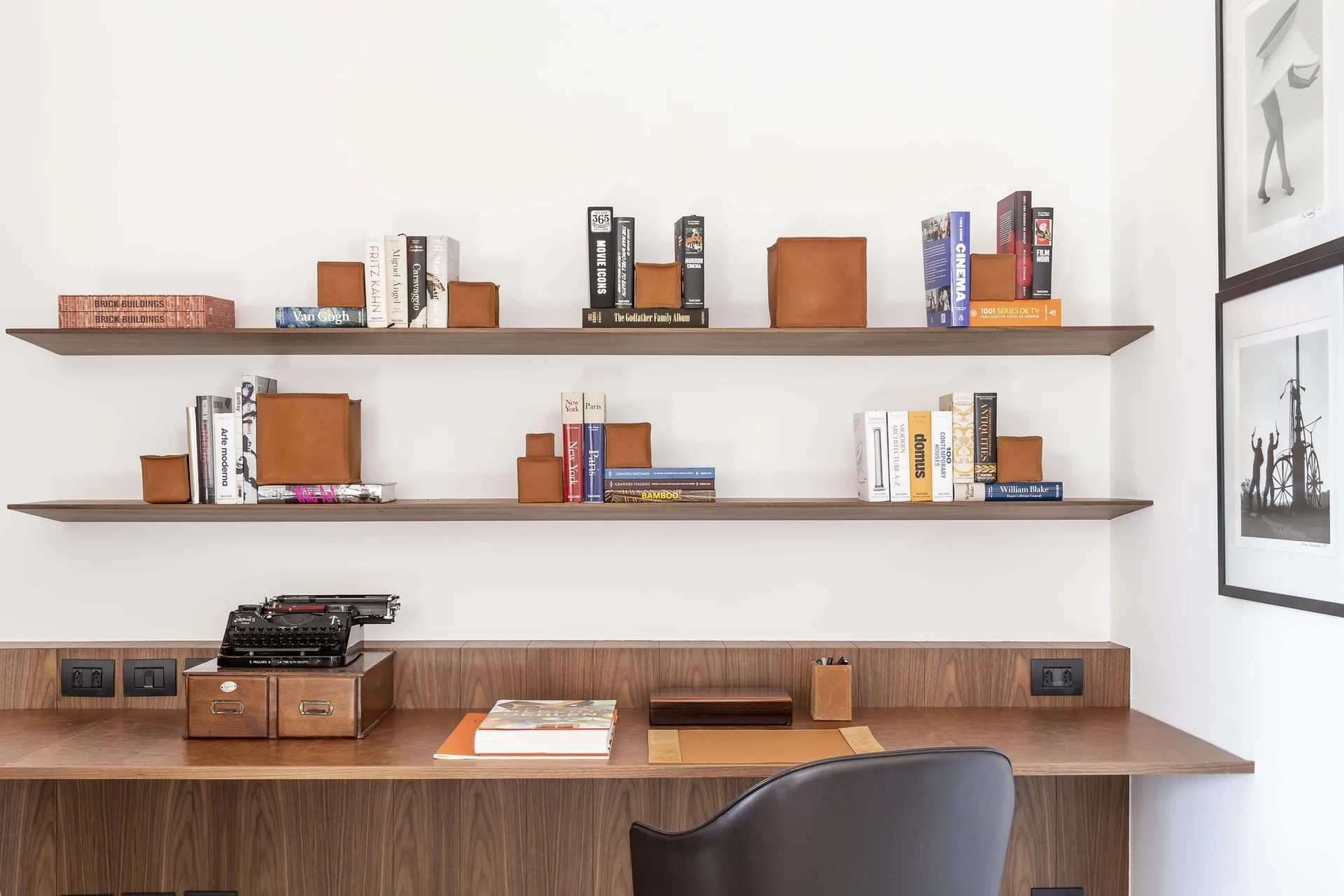 A built-in wooden desk features floating shelves styled with books and warm leather storage boxes next to black and white framed art.