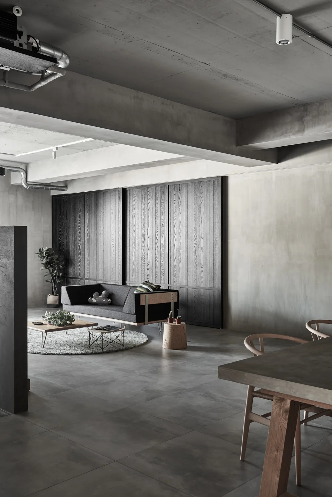 A minimalist living space featuring an exposed concrete ceiling and walls, a low gray sofa on a round rug, and dark wood sliding panels.