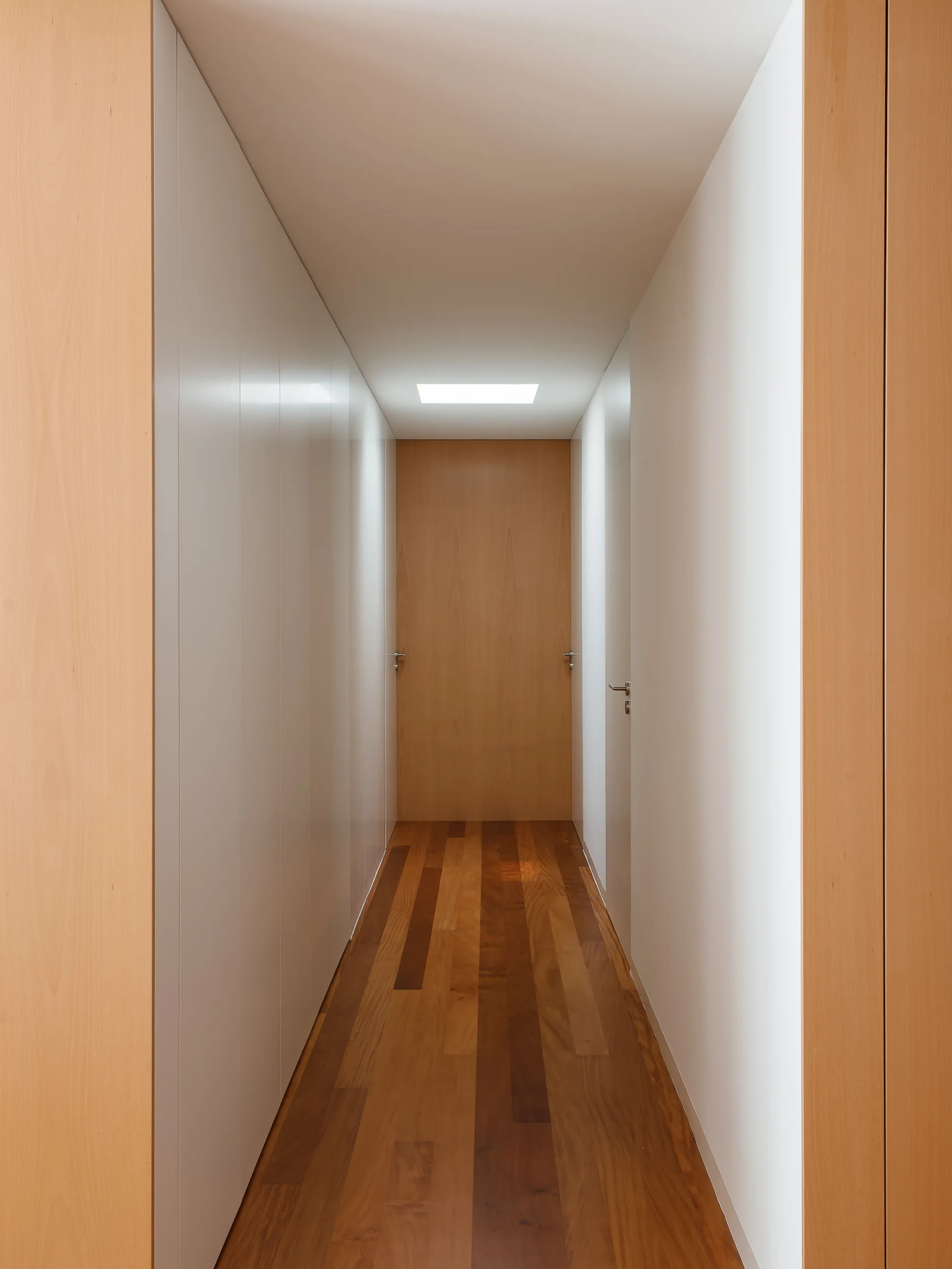 A narrow minimalist hallway features white flush wall panels, medium-toned wood plank flooring, and a wood door illuminated by a small skylight.