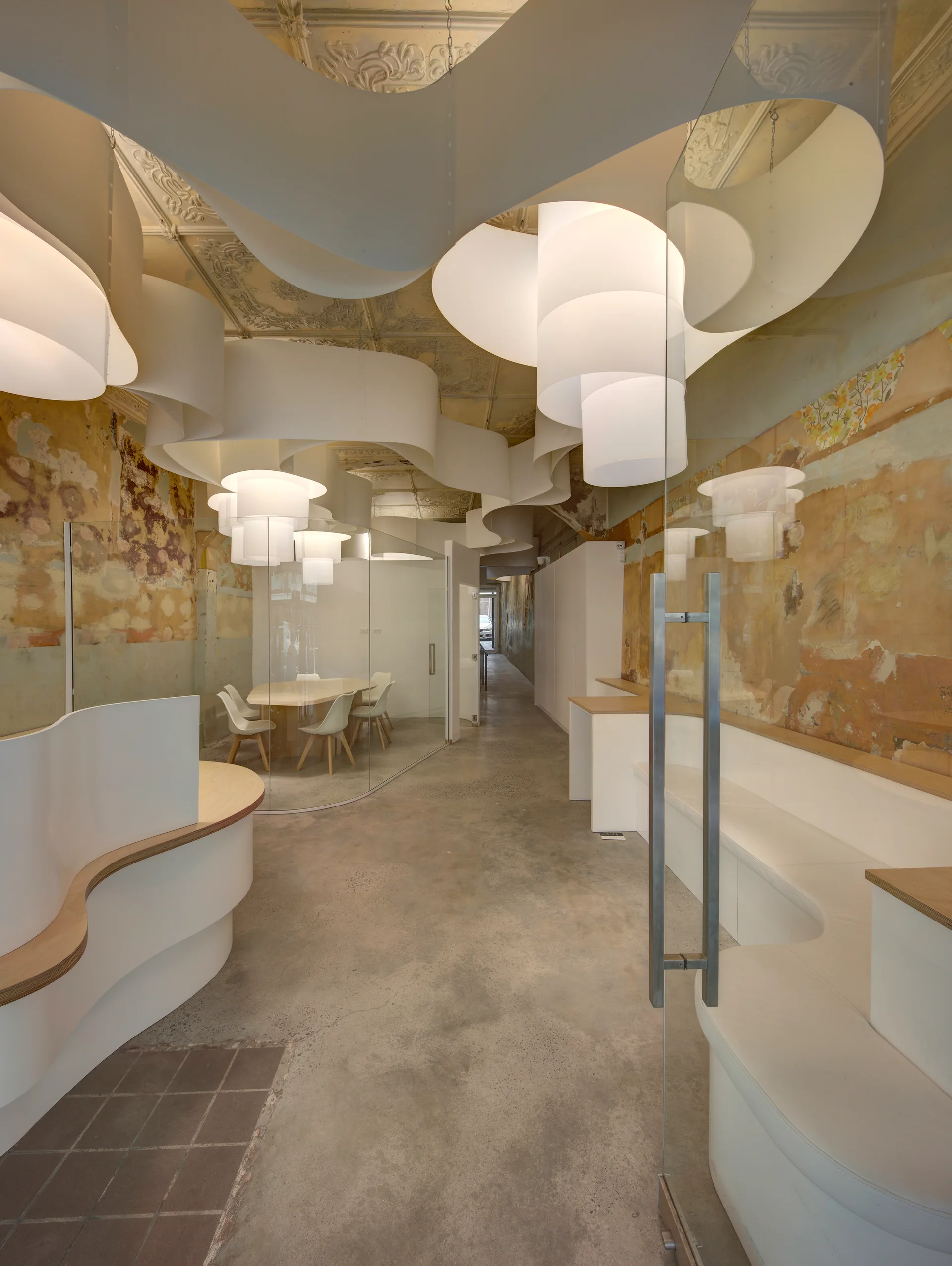 Modern office lobby with a flowing white ceiling installation, glass conference rooms, a curved reception desk, and distressed plaster walls.