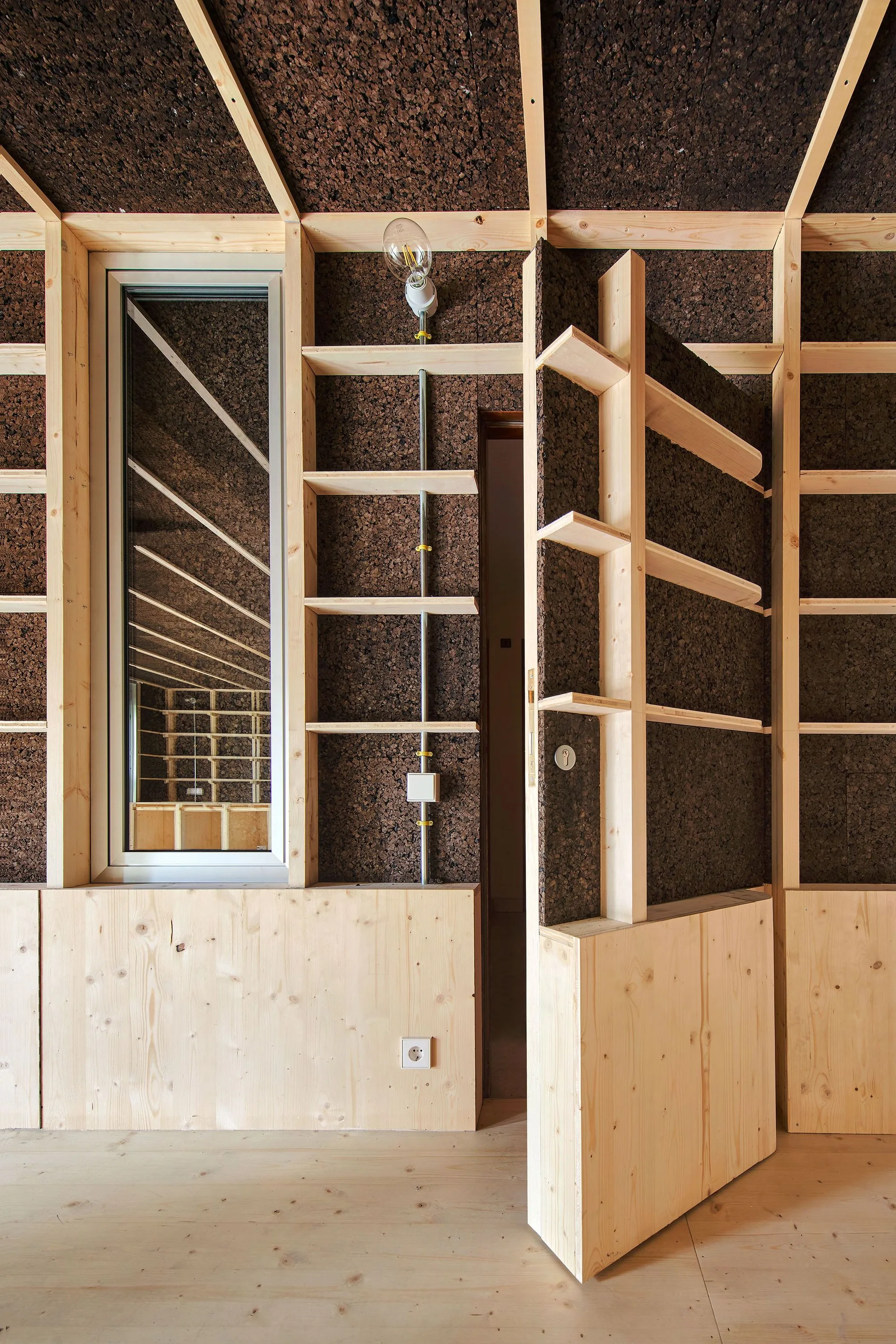 A modern interior wall features dark cork panels inside a light wood grid frame with integrated shelving and a matching open door.