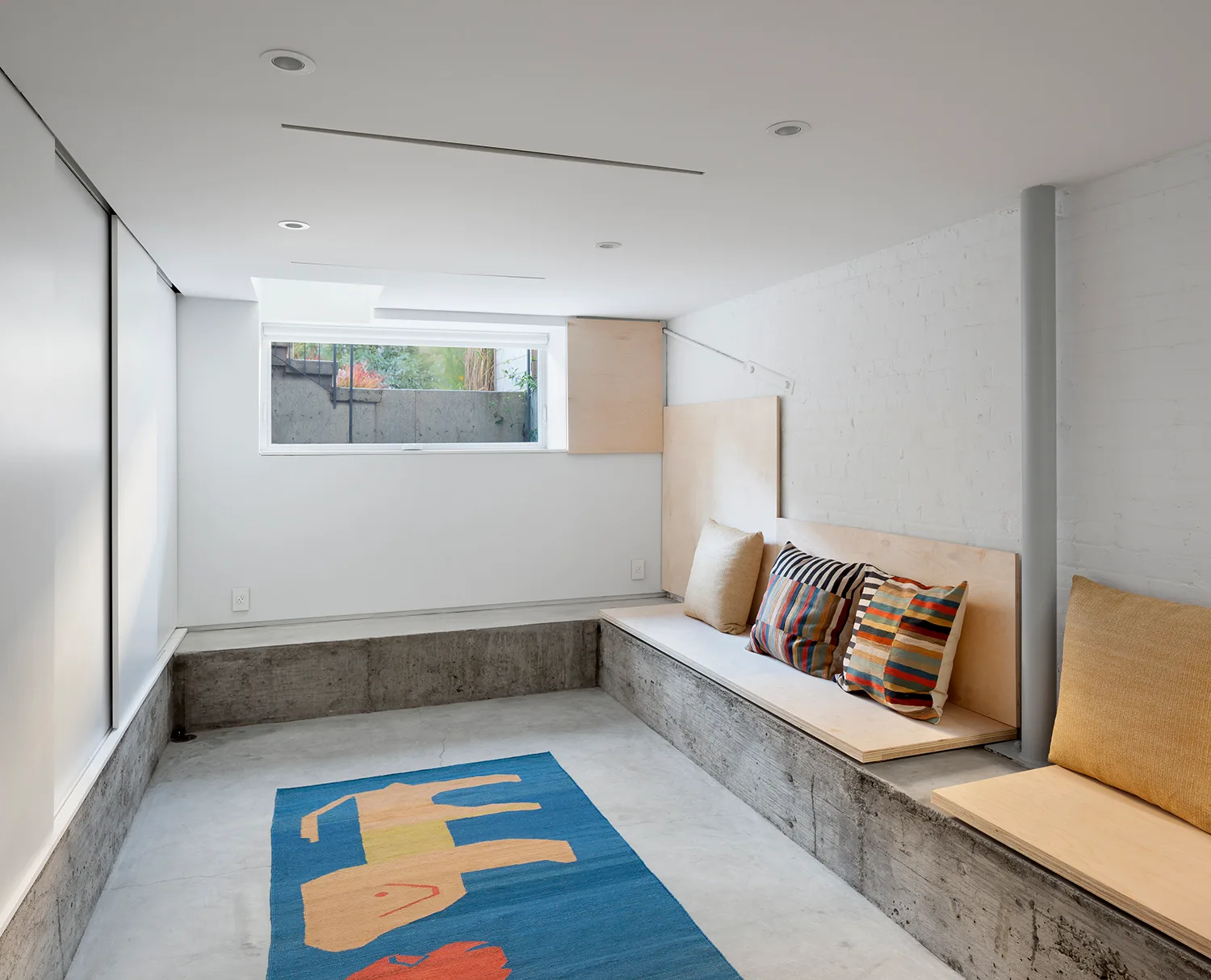 A minimalist basement room featuring a low concrete built-in bench topped with plywood and decorated with colorful patterned throw pillows.
