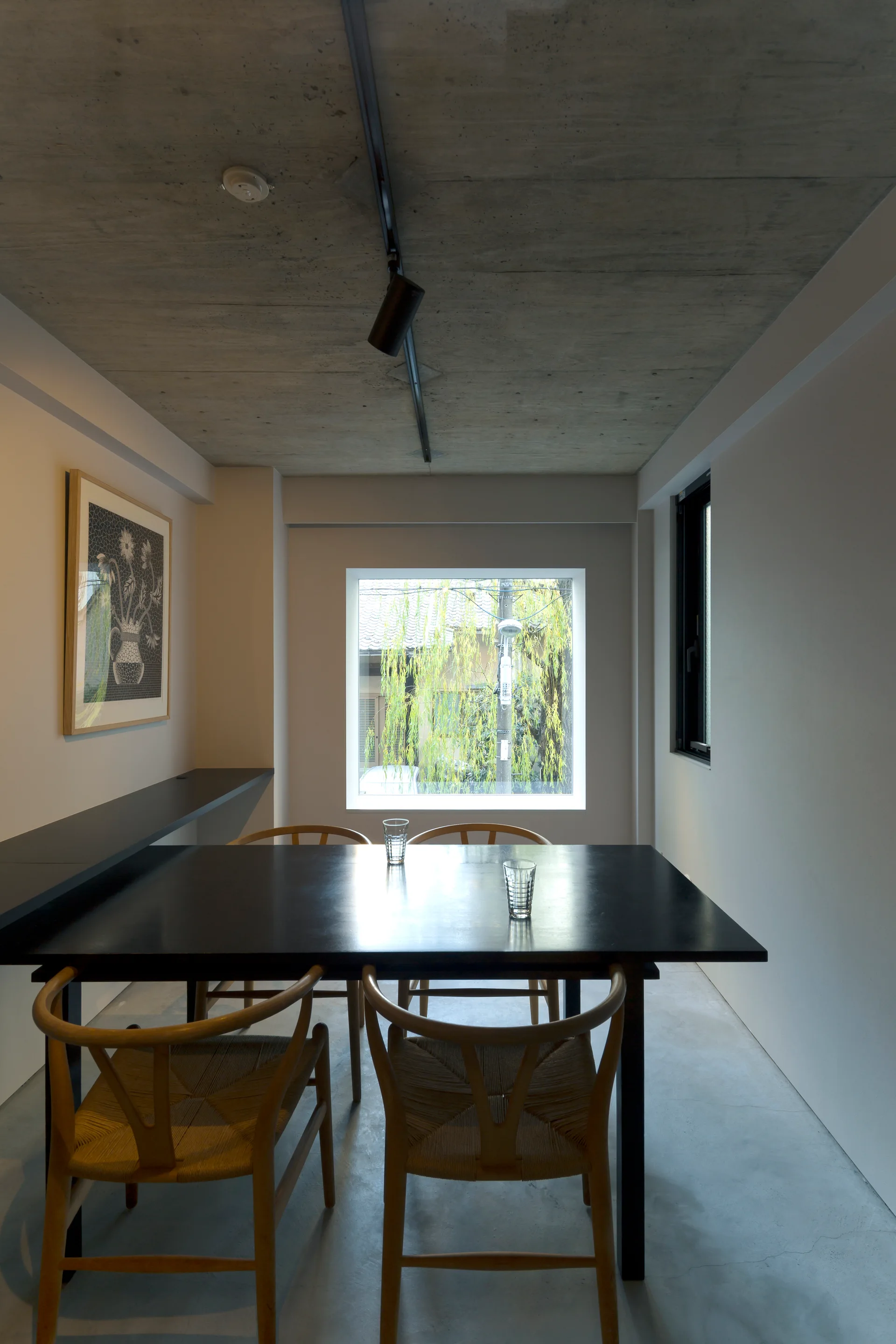 A minimalist dining room features a black table surrounded by light wood chairs under an exposed concrete ceiling with a square window in the background.