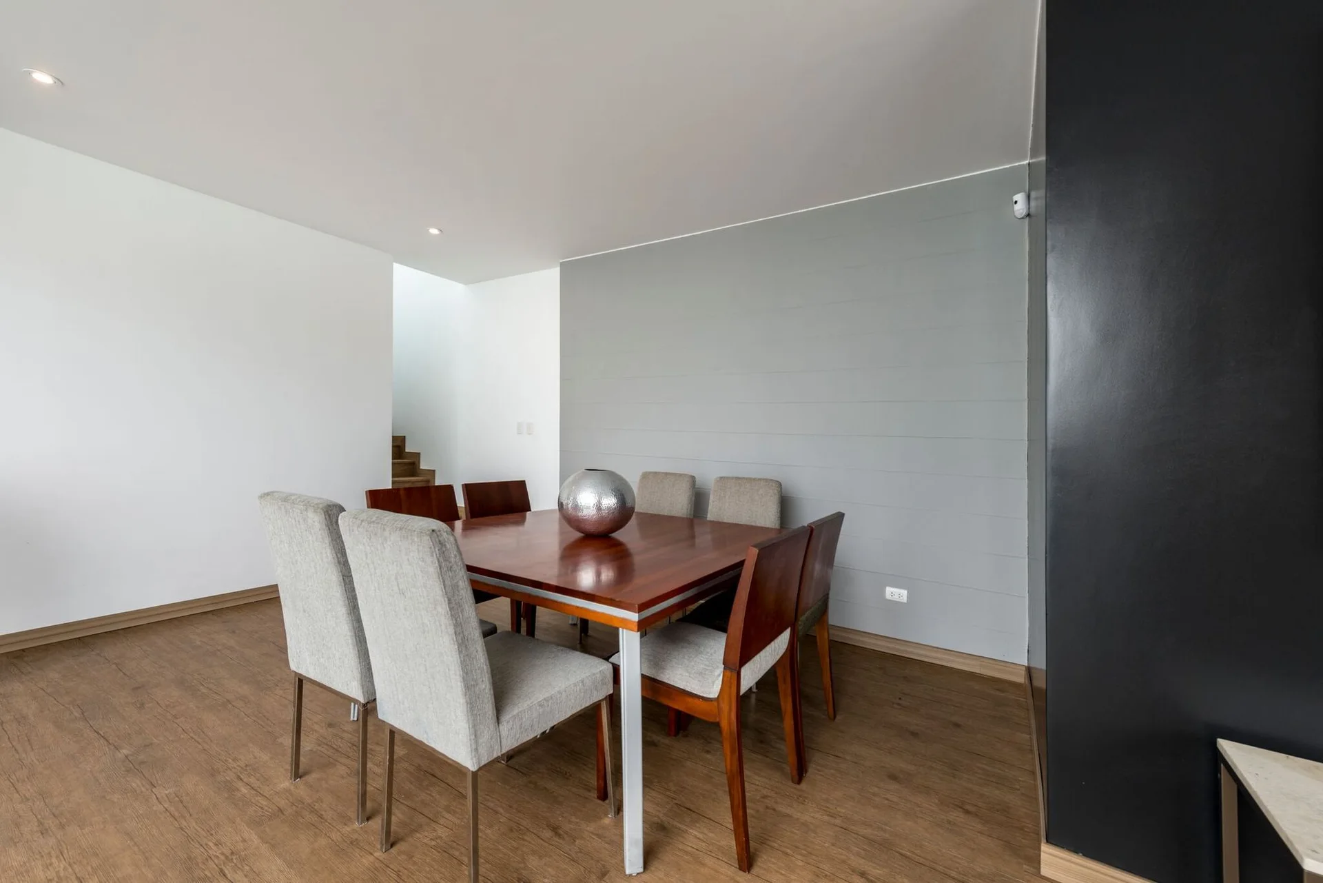 A square wood dining table with mixed chairs sits on wood flooring in front of a grey horizontally paneled accent wall.
