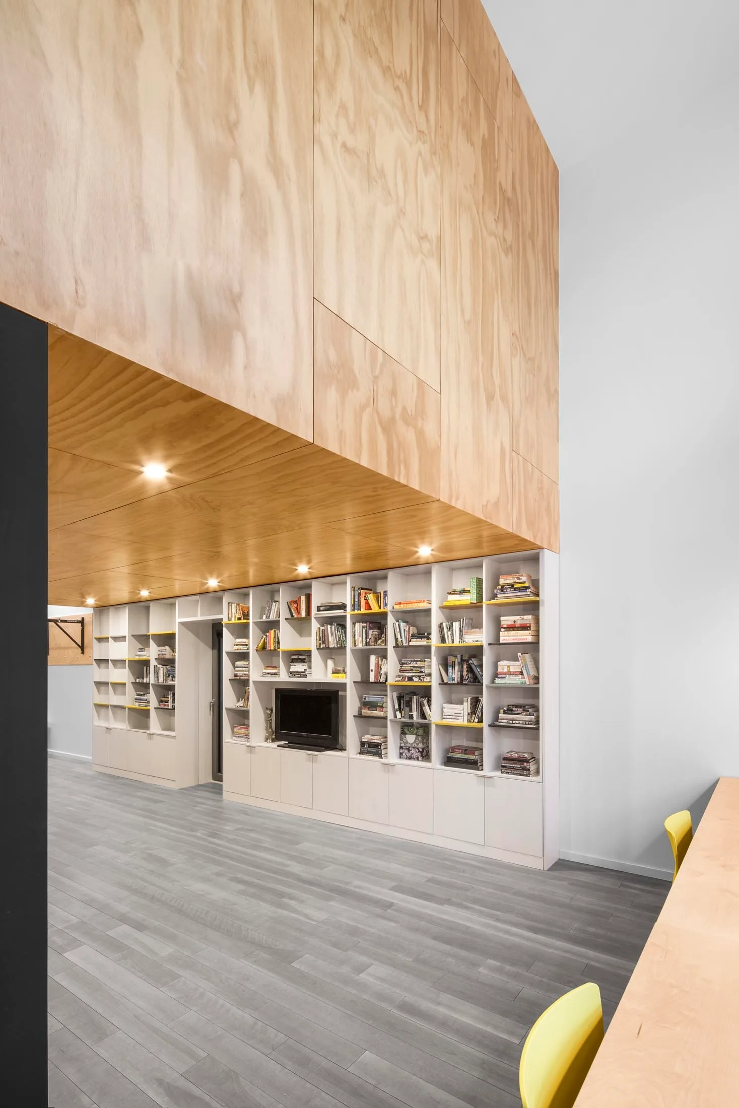 A modern interior features a large plywood paneled ceiling canopy positioned above white built-in bookshelves filled with books.