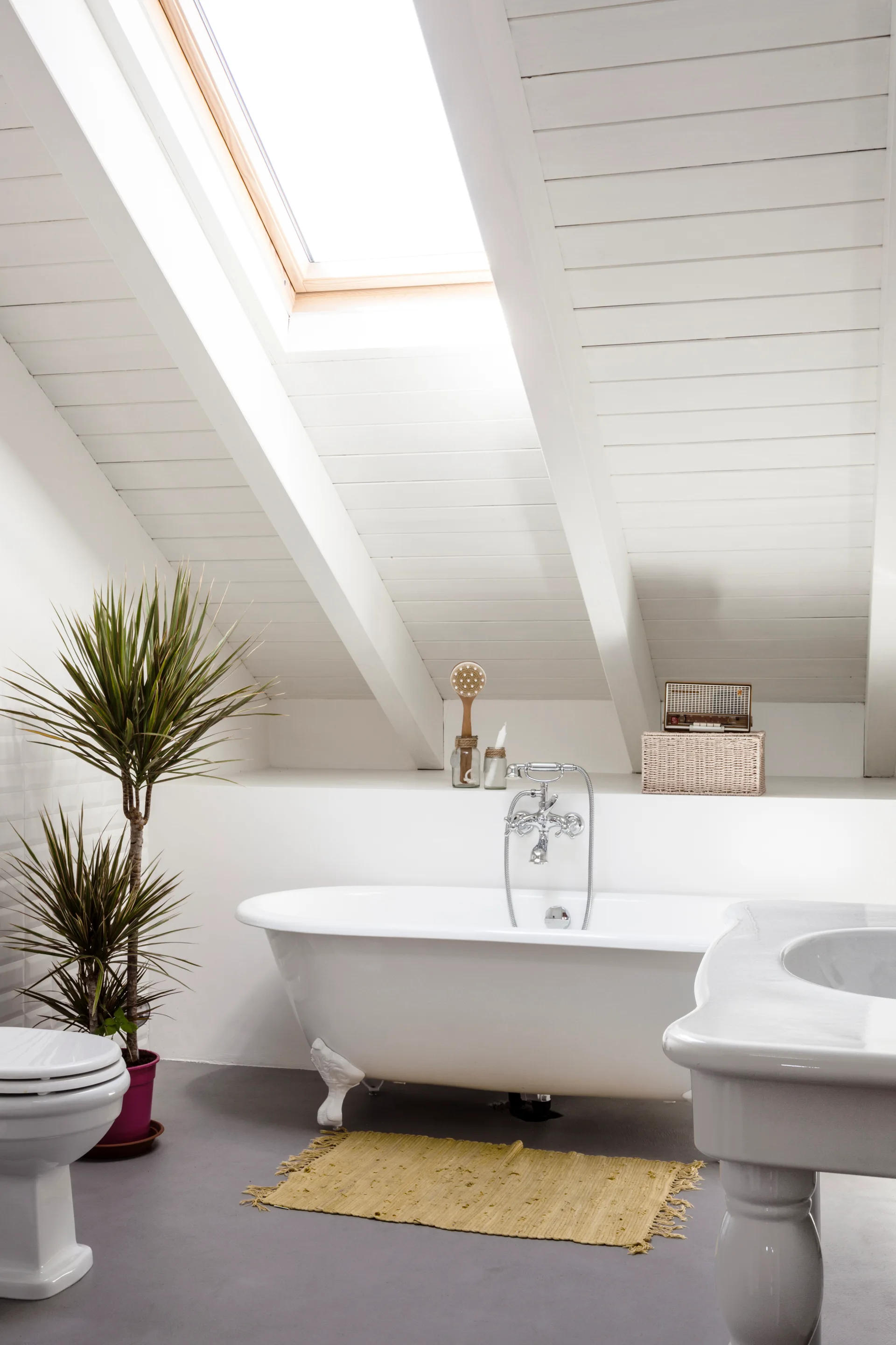 A bright attic bathroom featuring a white clawfoot tub under a skylight in a slanted wood plank ceiling, alongside a vanity and toilet.