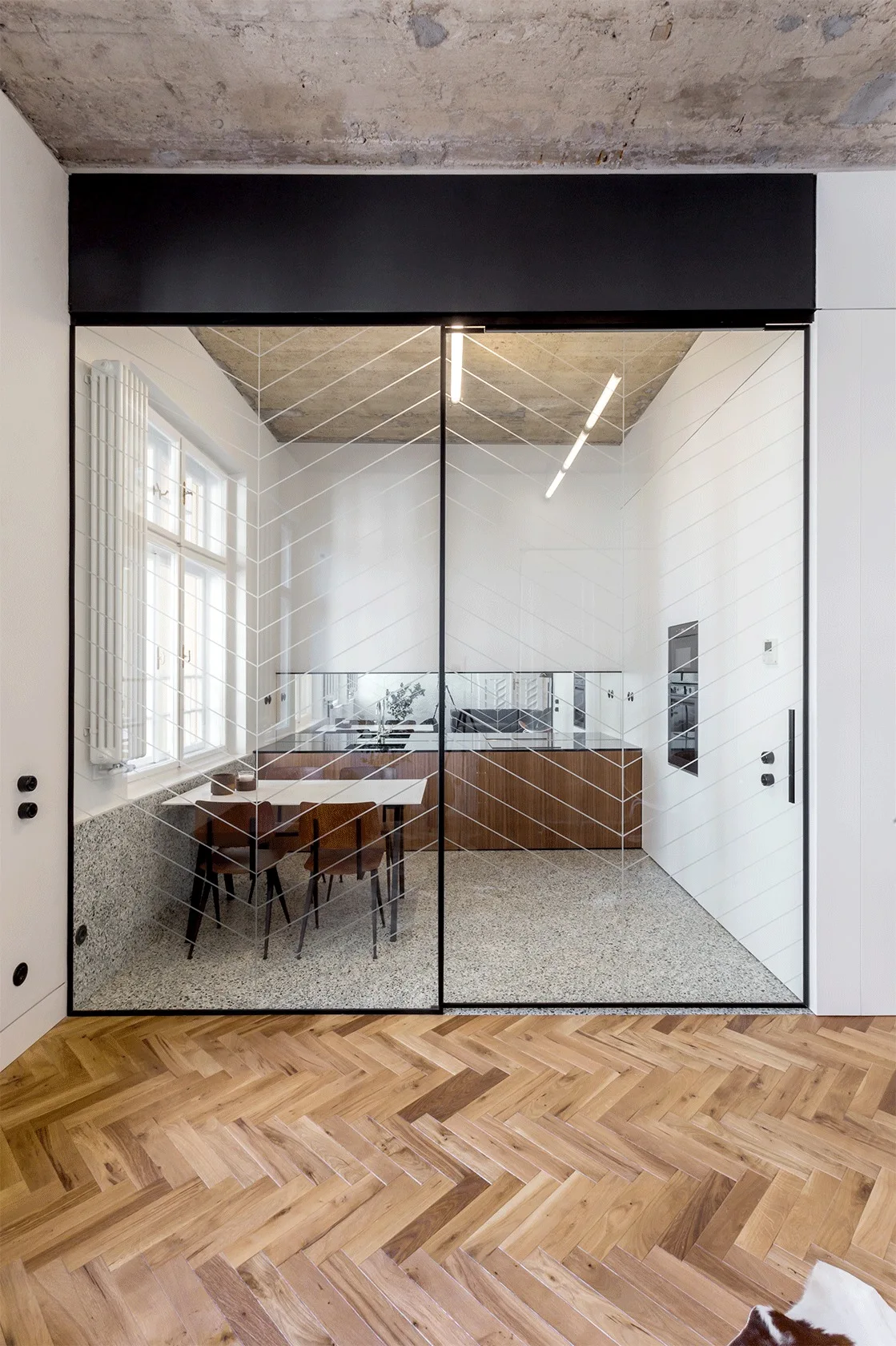 A modern kitchen and dining space featuring terrazzo floors and wood cabinetry, separated by a glass partition with a white chevron pattern.
