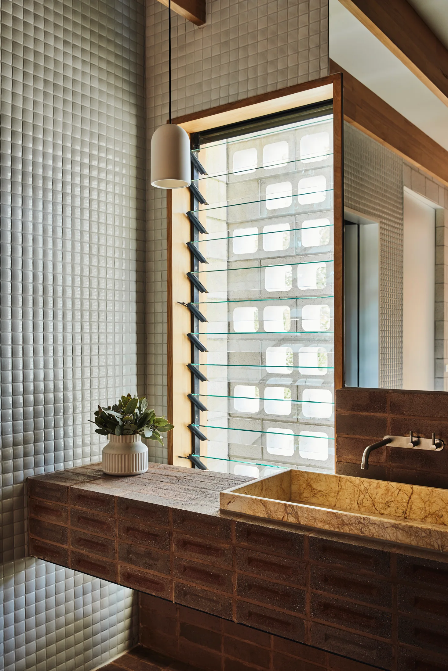 A modern bathroom features a cantilevered brick vanity with a stone basin sink next to a wood-framed glass louvre window and a tiled wall.