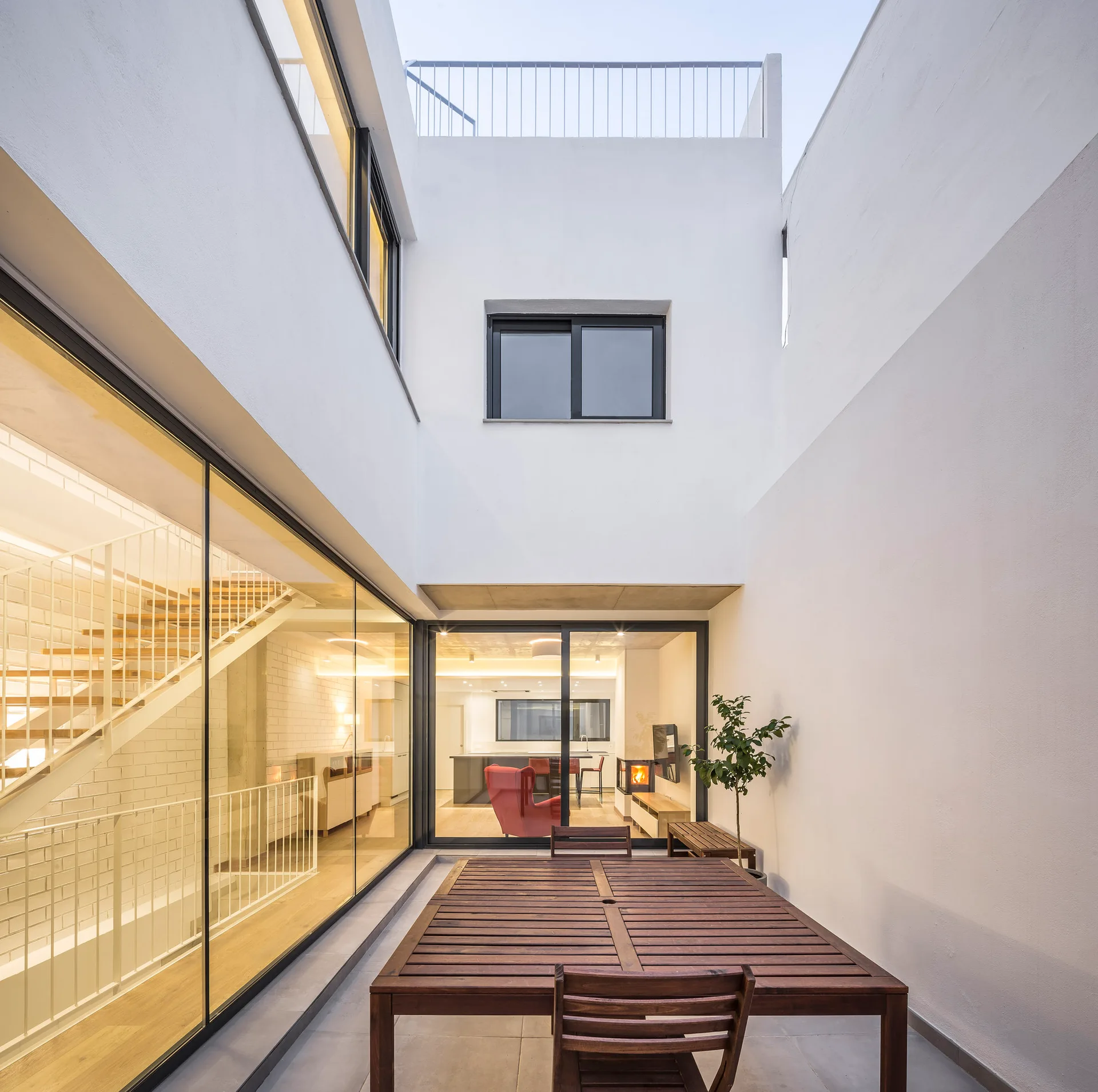 A modern enclosed courtyard features a wooden dining table and chairs, surrounded by white stucco walls and large glass doors looking inside.