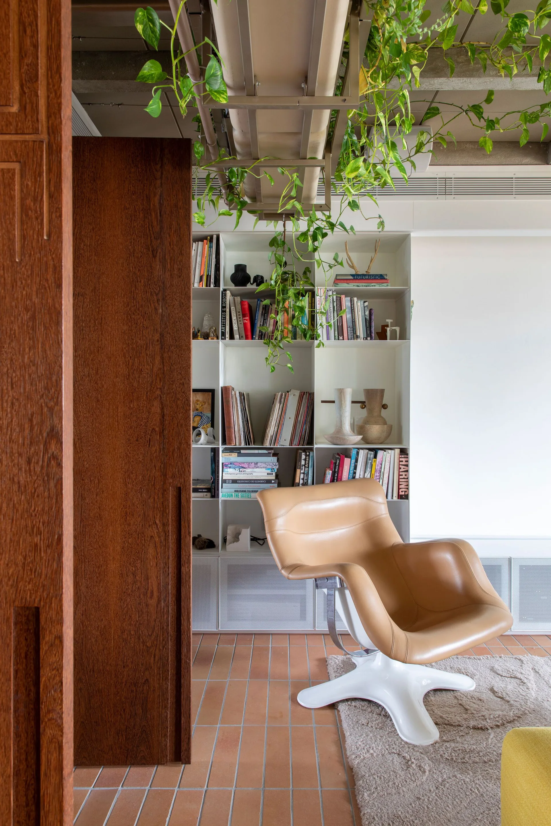 A tan leather lounge chair sits on a shag rug in front of a white bookshelf, with a trailing green plant hanging from the exposed ceiling.
