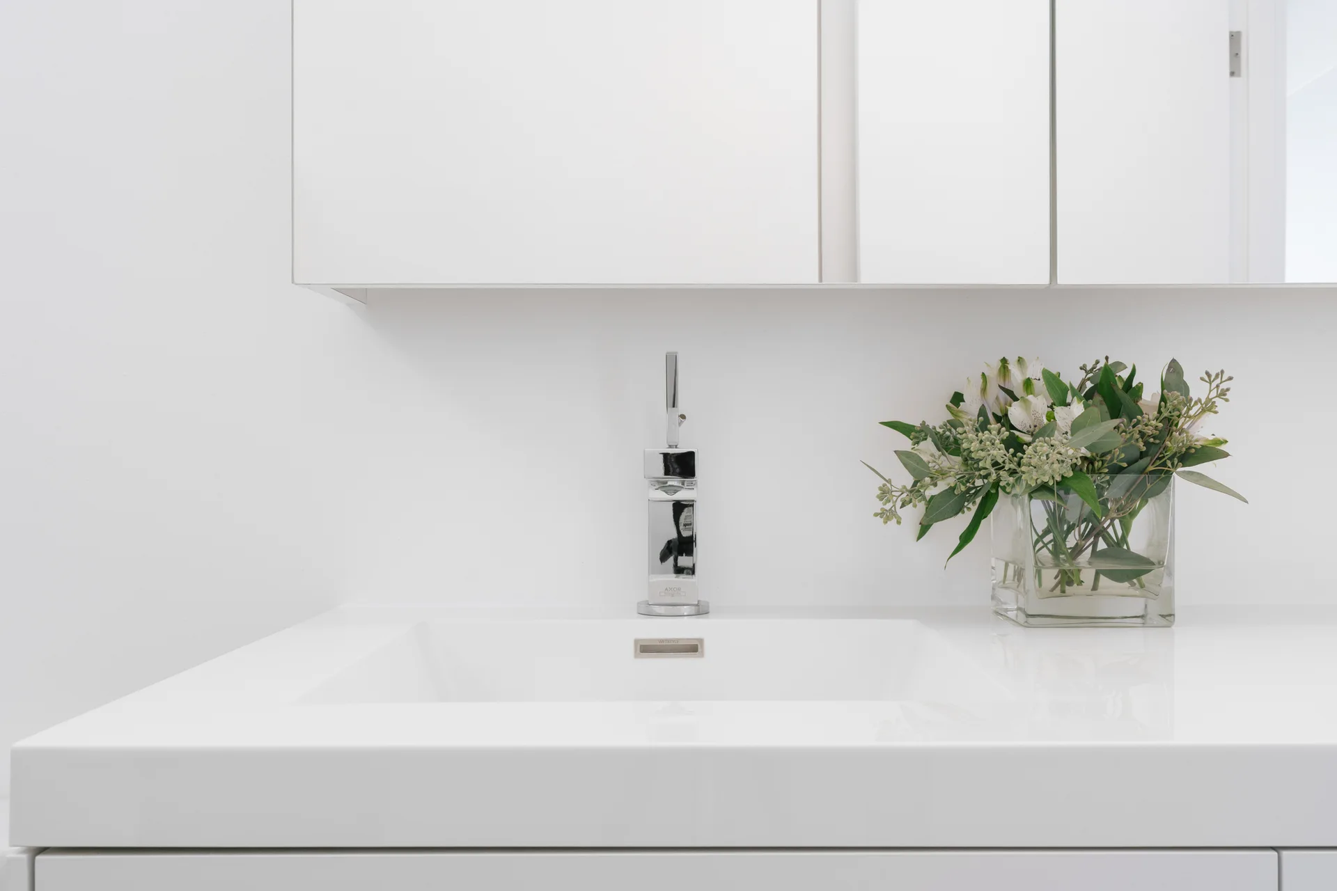 A minimalist white bathroom vanity features an integrated countertop sink, a polished chrome faucet, and a glass vase with greenery.