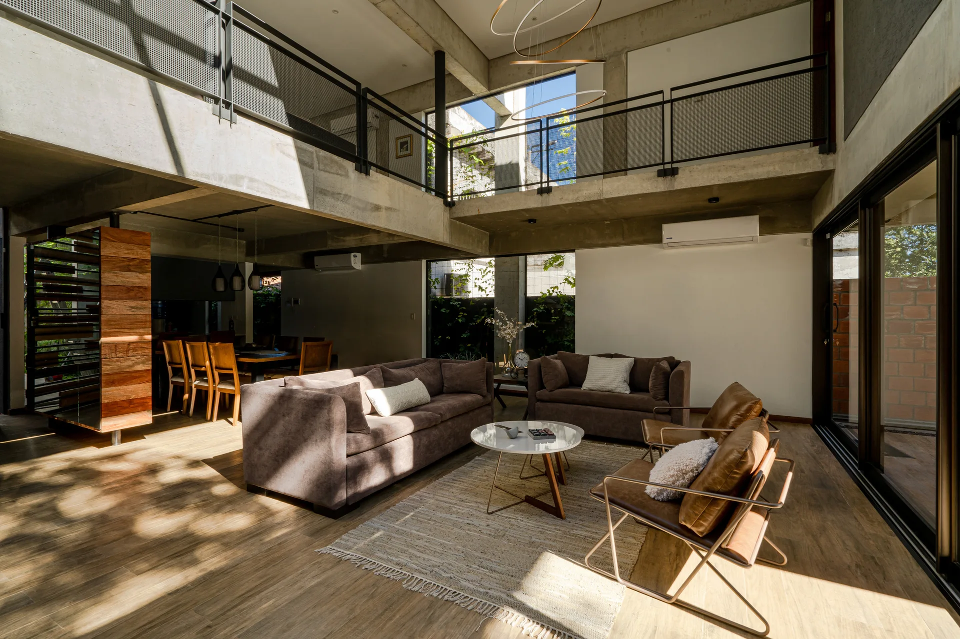 An industrial living room features brown sofas, tan leather chairs, and a round coffee table beneath a concrete mezzanine and exposed beams.