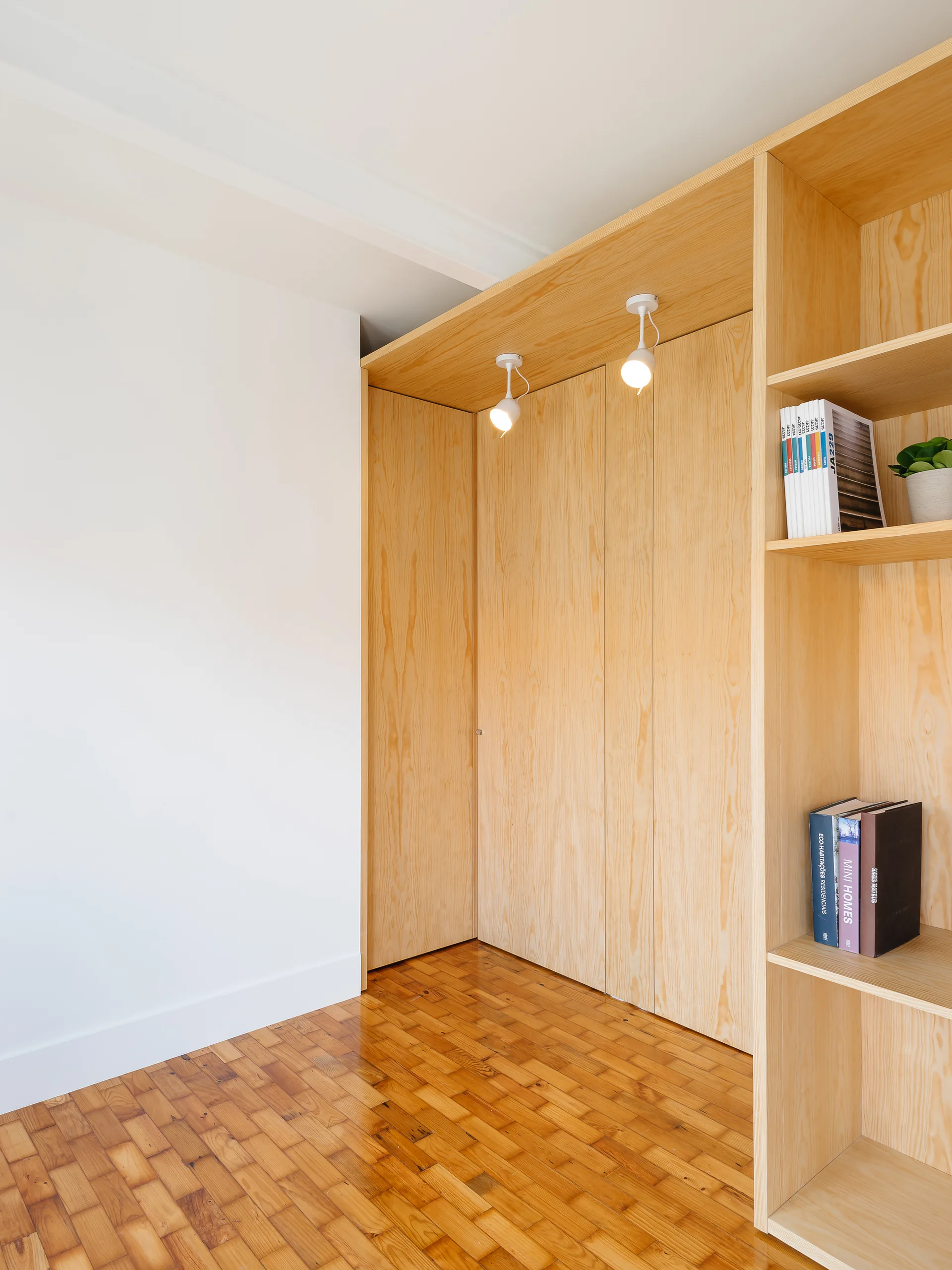 A light wood built-in closet with open shelves and overhead spotlights sits on a parquet wood floor next to a white wall.
