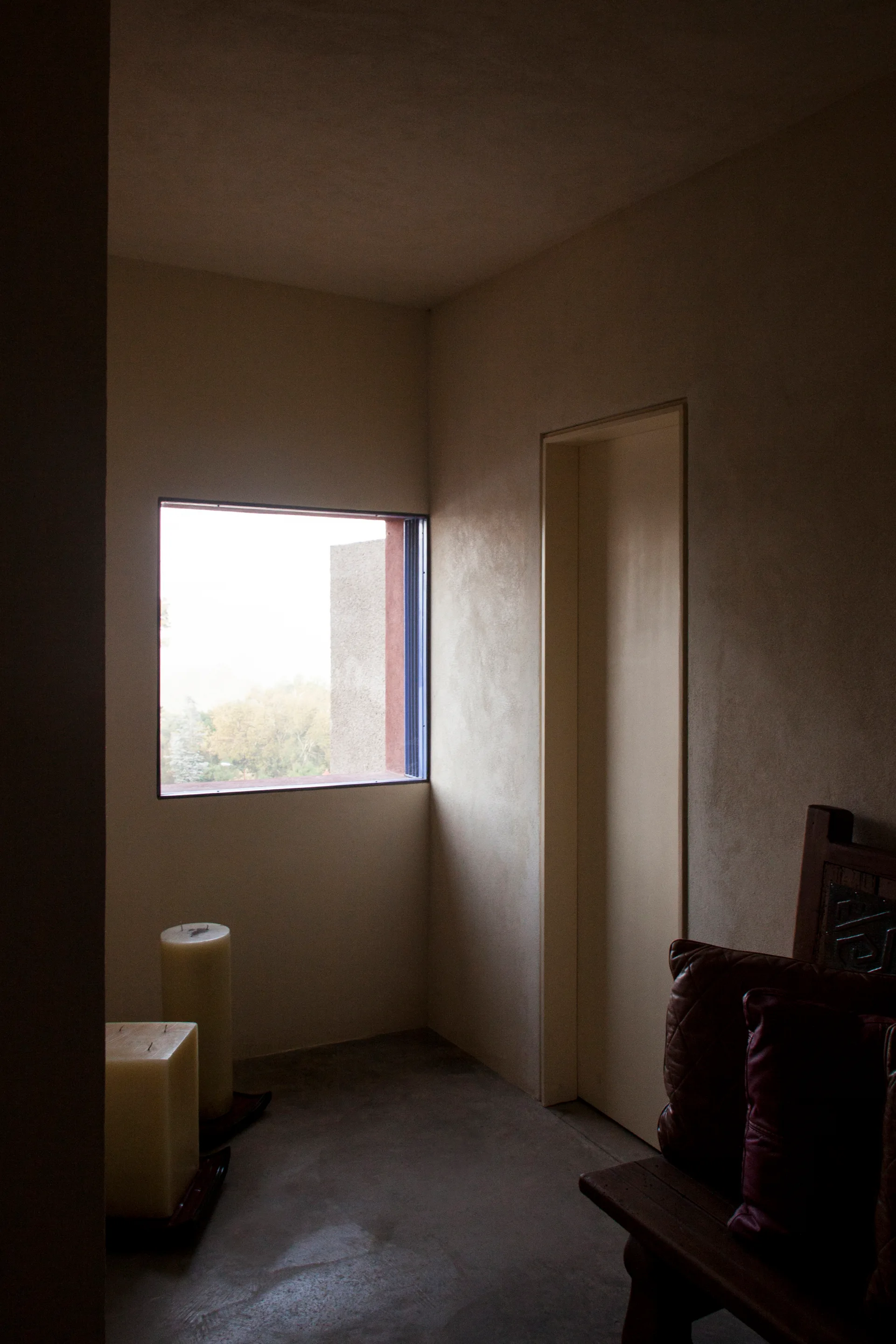 A dim minimalist room corner with textured beige plaster walls, a recessed square window, and a dark wooden bench with leather cushions.