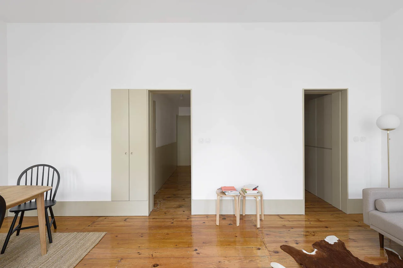 A minimalist white room features polished wood floors, two open doorways with painted beige trim, a small dining table, and a grey sofa.