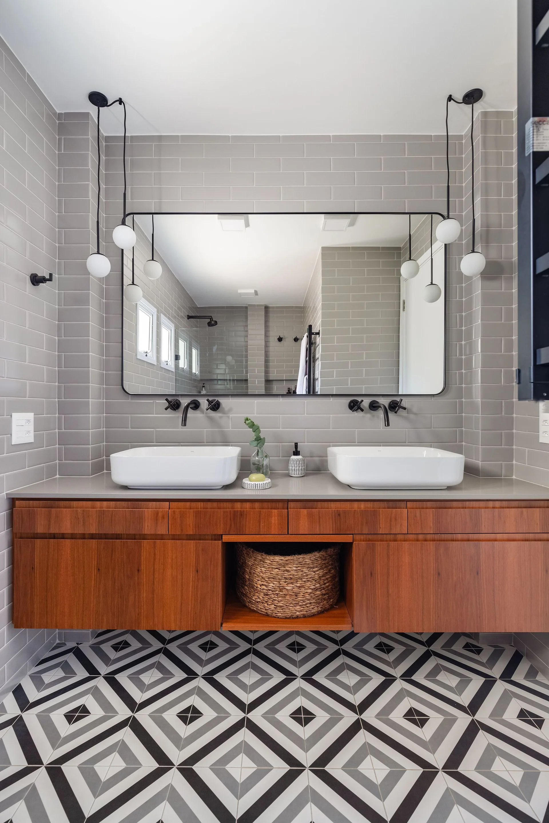 A contemporary bathroom with a floating wood vanity, white vessel sinks, gray subway tile walls, and geometric patterned floor tile.
