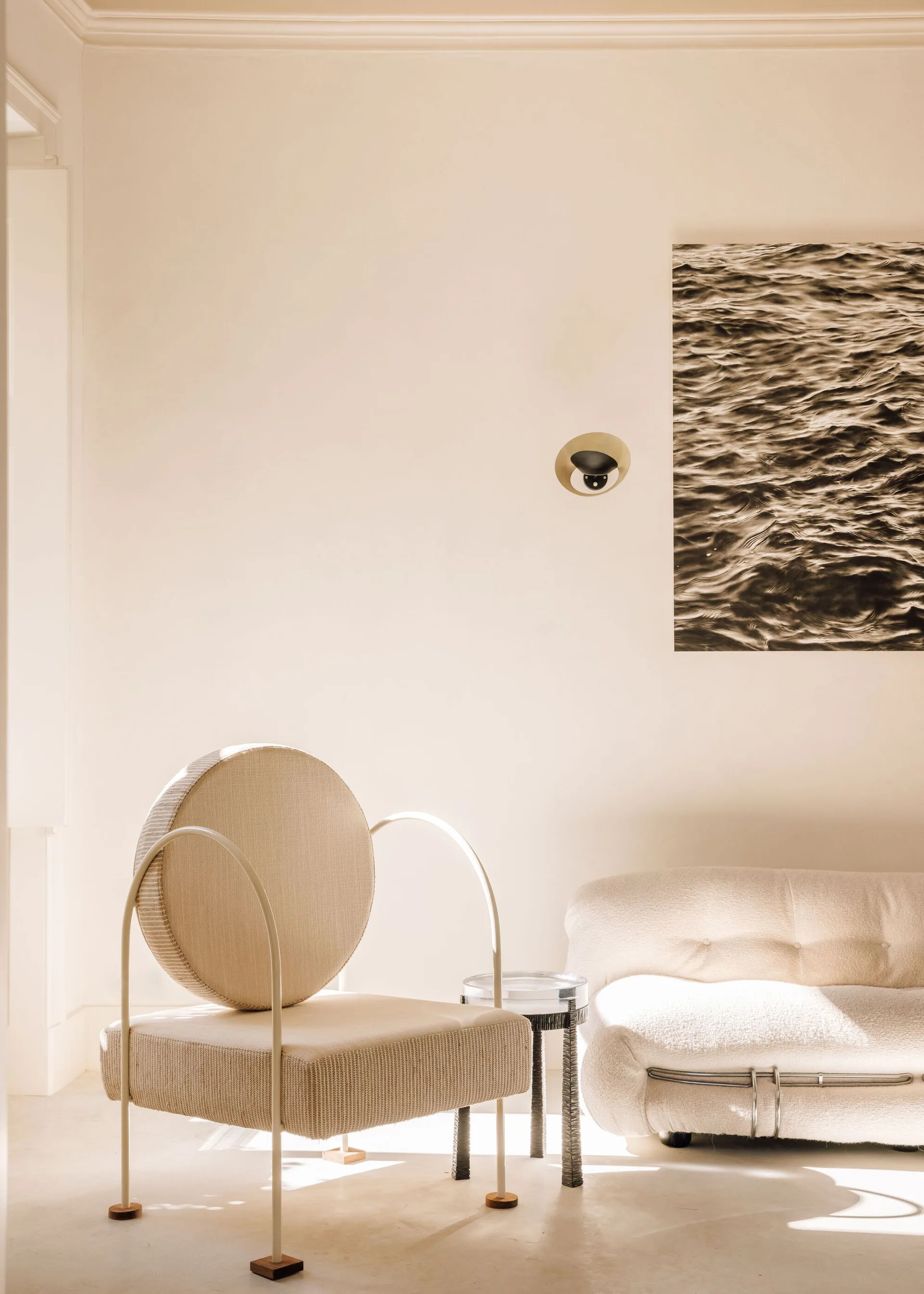 A minimalist living room features a cream armchair with a circular backrest, a low tufted sofa, and a black and white photograph on the wall.