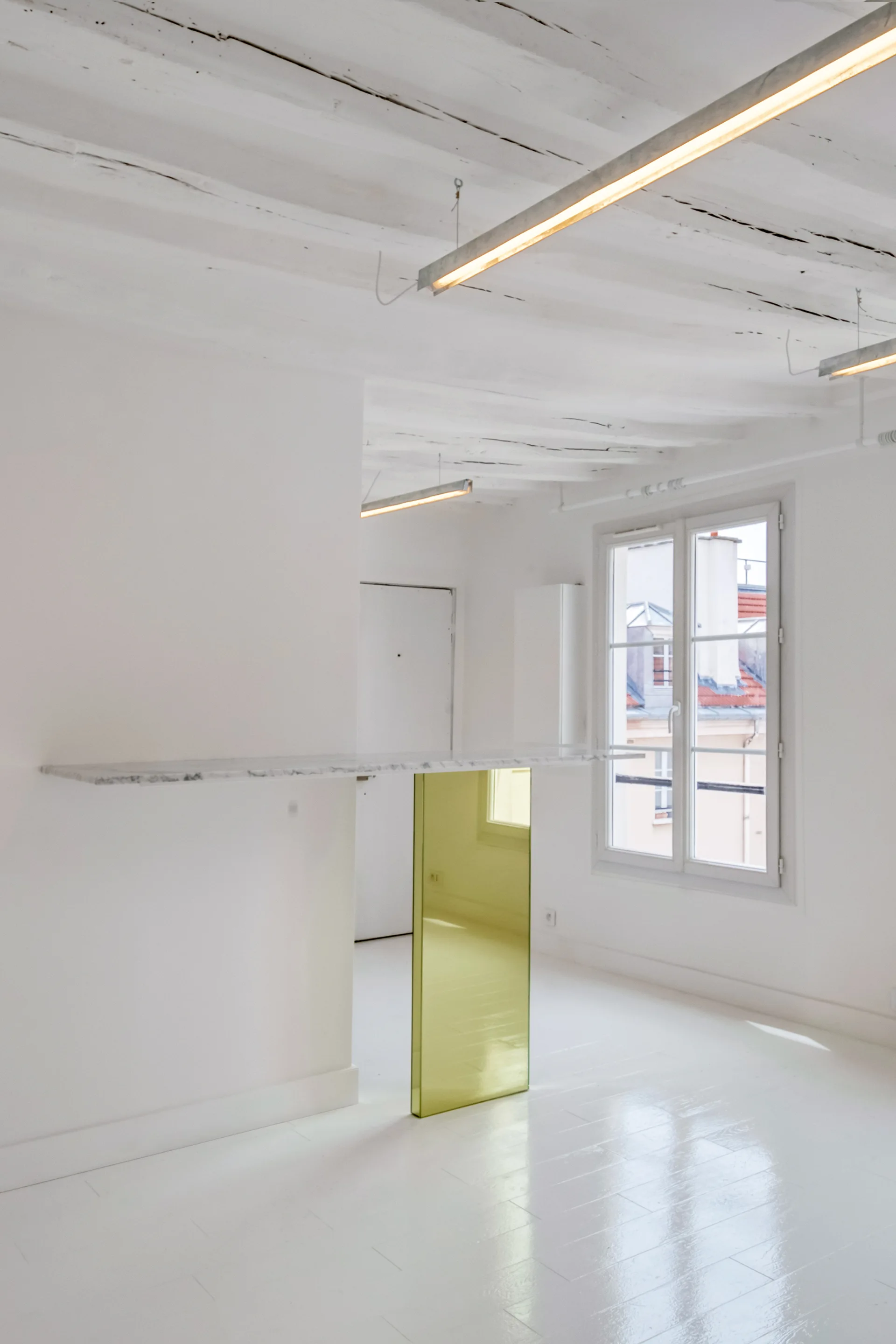 An all-white room featuring a floating marble console supported by a yellow glass panel, white painted ceiling beams, and glossy floors.
