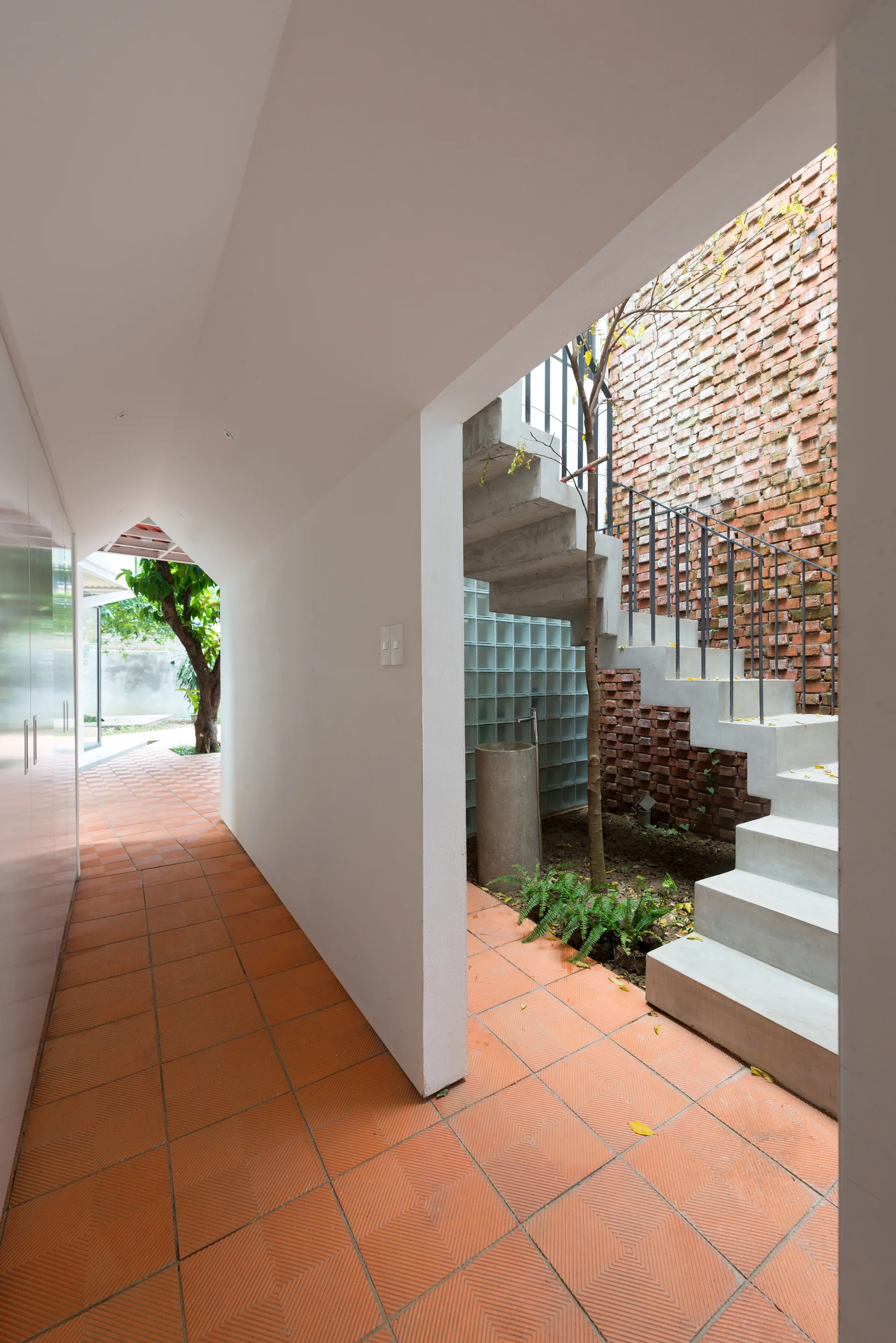 A hallway with terracotta tile flooring opens to an outdoor courtyard featuring a raw concrete staircase against an exposed red brick wall.