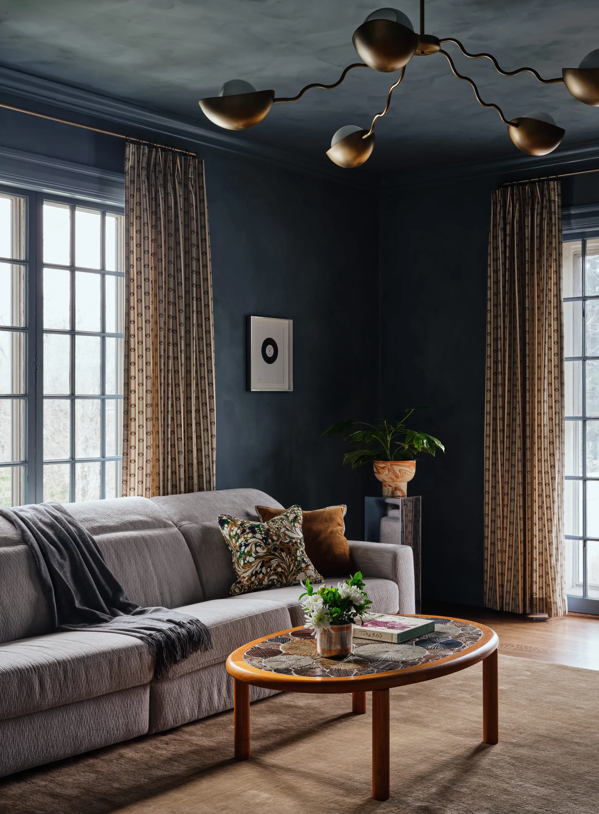 A moody living room features dark navy blue walls and ceiling, a textured light grey sofa, a patterned oval coffee table, and a brass chandelier.