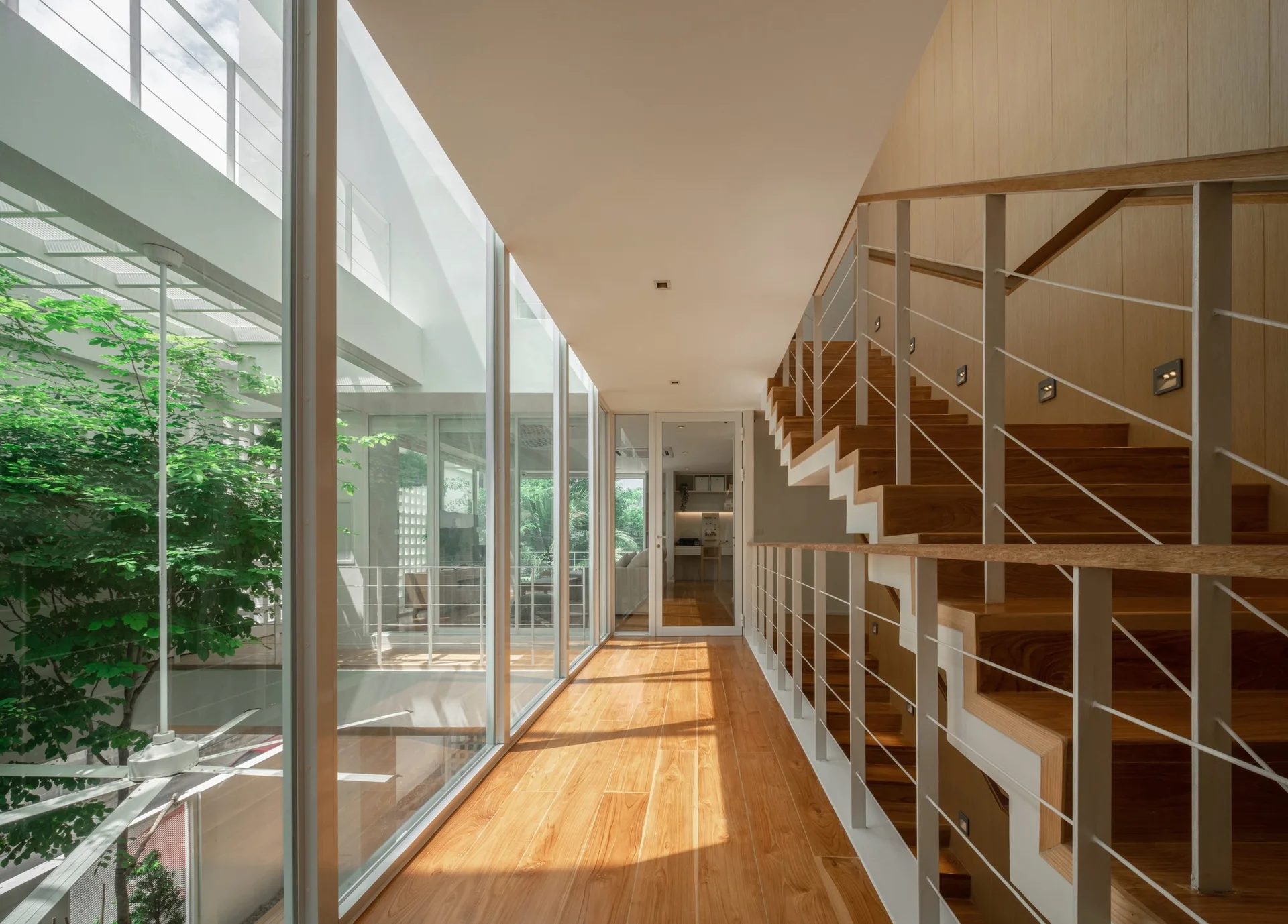A wood-floored hallway features a modern staircase with thin white railings opposite floor-to-ceiling glass walls looking out to a leafy courtyard.