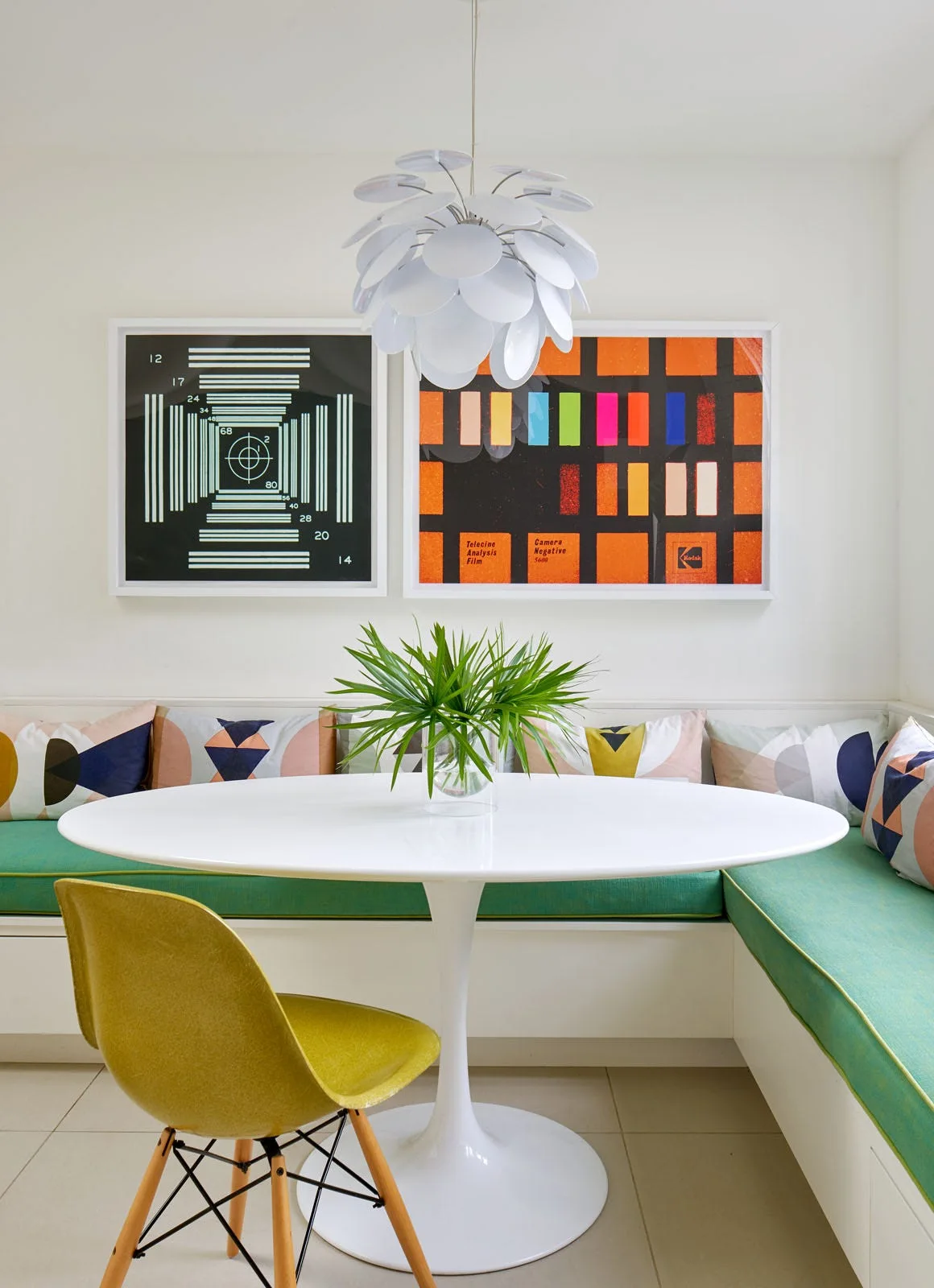 A modern dining nook features a white tulip table, built-in corner bench with green cushions, a yellow chair, and framed graphic art.