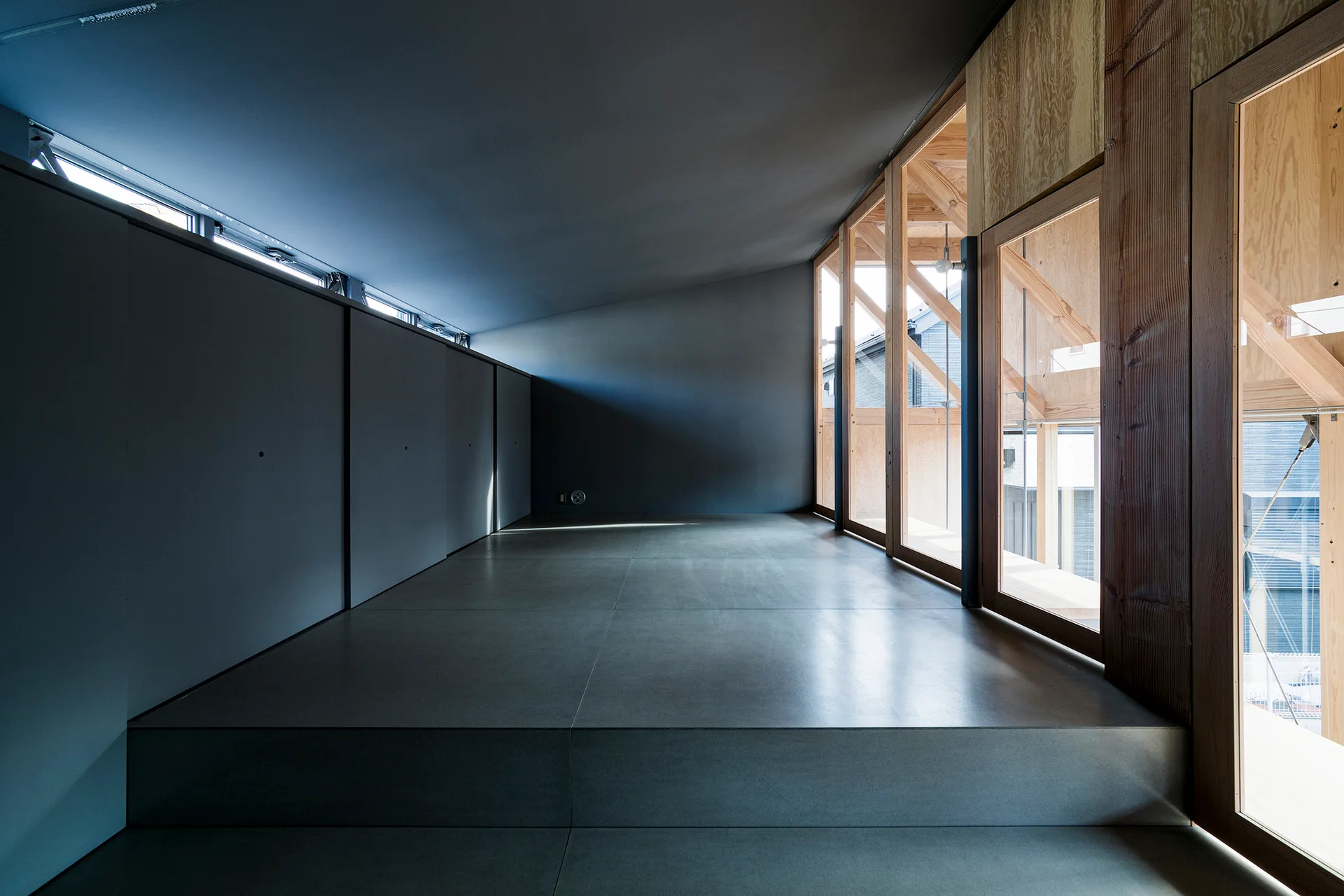 A minimalist hallway with matte charcoal grey walls and floor panels contrasting with exposed timber-framed glass windows on the opposite side.
